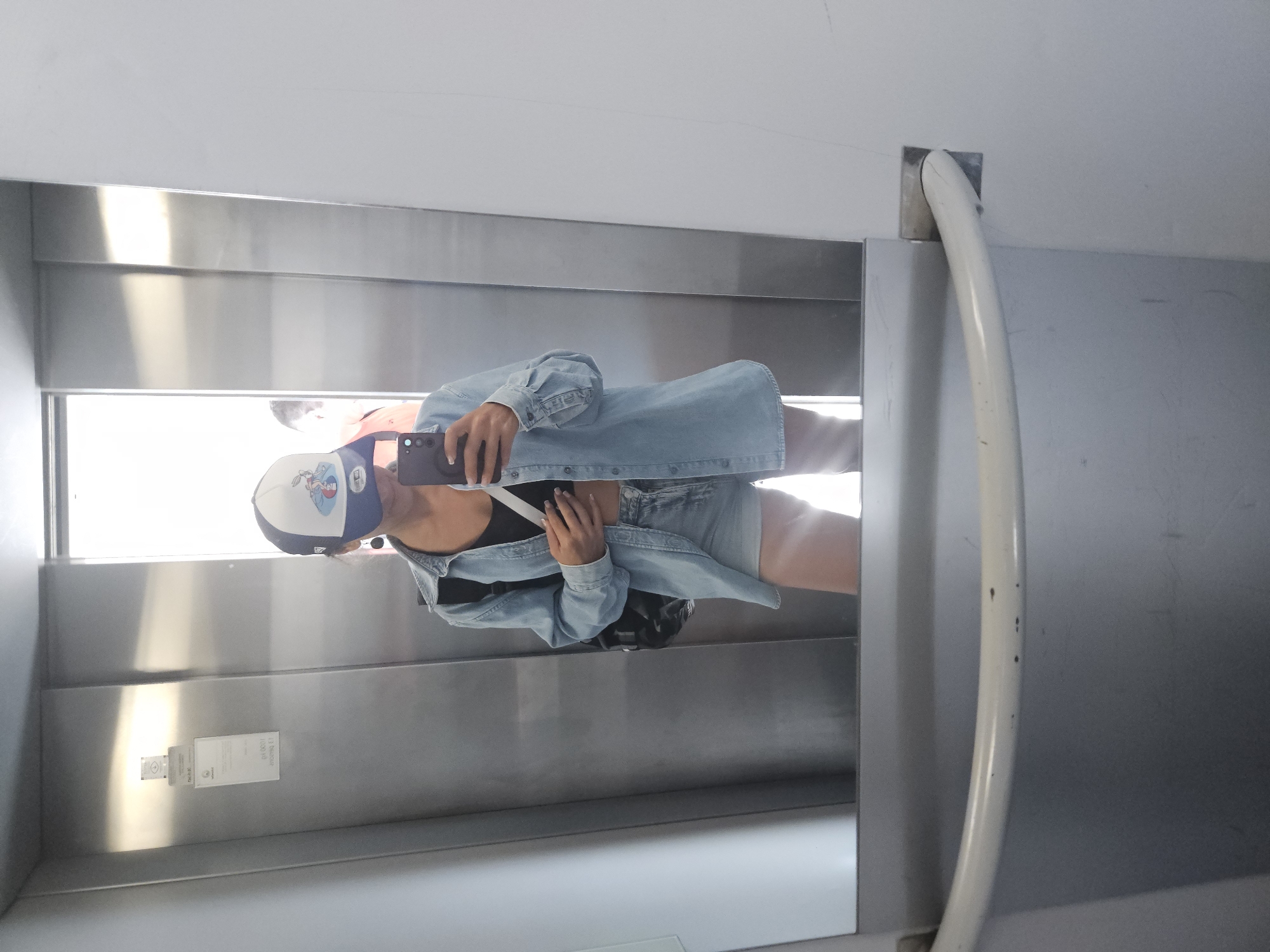 A person wearing a denim jacket, denim shorts, a black crop top, and a baseball cap is taking a selfie in a mirror. They are holding a smartphone in their right hand and their left hand is resting on their stomach. A backpack is visible on their left shoulder. The background appears to be an elevator with metallic walls and a white door frame. The scene is lit by artificial light. Based on the visible text in the image, it's possible the location is an elevator in Budva, Montenegro, given the presence of Cyrillic and Latin script on a sign attached to the wall, which includes text that is partially obscured but suggests a European location. The mirror reflects the person and the elevator interior, with a slight glare from the lighting.