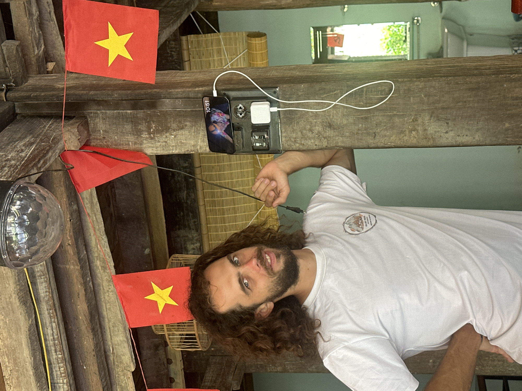 The image displays a young man in an indoor or covered outdoor setting characterized by rustic wooden beams, suggesting a guesthouse or traditional home in Du Già Commune, Vietnam. The man, with long, curly brown hair, a beard, and light-colored eyes, looks directly at the camera with a neutral to slightly smirking expression. He is wearing a plain white short-sleeved t-shirt with an indistinct circular logo on the chest. His right arm is bent, holding a black charging cable.
Above and to the left of the man (when viewed upright), small red Vietnamese flags with a yellow star are strung along a wooden beam, indicating the location. A clear, faceted light fixture, resembling a small disco ball, hangs on the far left.
Mounted on one of the horizontal wooden beams is a black charging station. A dark-colored smartphone, displaying the time "10:51" and a colorful wallpaper, rests on this station, connected by a white cable to a white power adapter plugged into one of the outlets. Another white cable loops loosely on the beam. In the background, woven bamboo or rattan mats are visible, contributing to the rustic aesthetic of the environment. The scene captures a moment of charging devices within a locally themed setting.