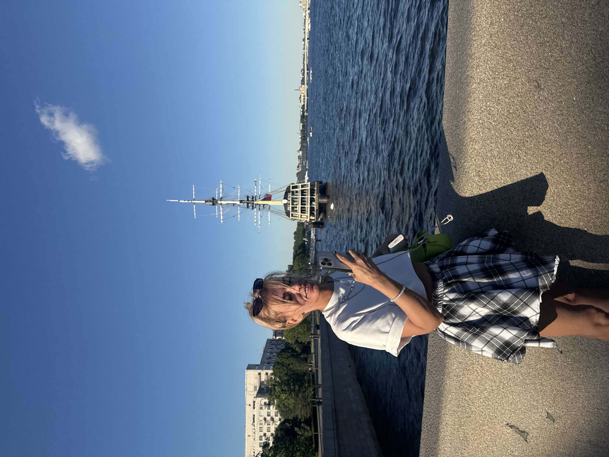 A young blonde woman with sunglasses perched on her head stands on a stone embankment, holding a smartphone. She is wearing a white crop top and a black and white plaid skirt. A green bag is tucked into her waist. In the background, a large sailing ship with multiple masts is visible on a body of water, likely the Neva River. The cityscape of Saint Petersburg, Russia, can be seen along the riverbank, with buildings and trees lining the waterfront. The sky is a clear blue with a single white cloud. The scene appears to be during daylight hours, possibly late afternoon or early evening, given the long shadows cast on the embankment.