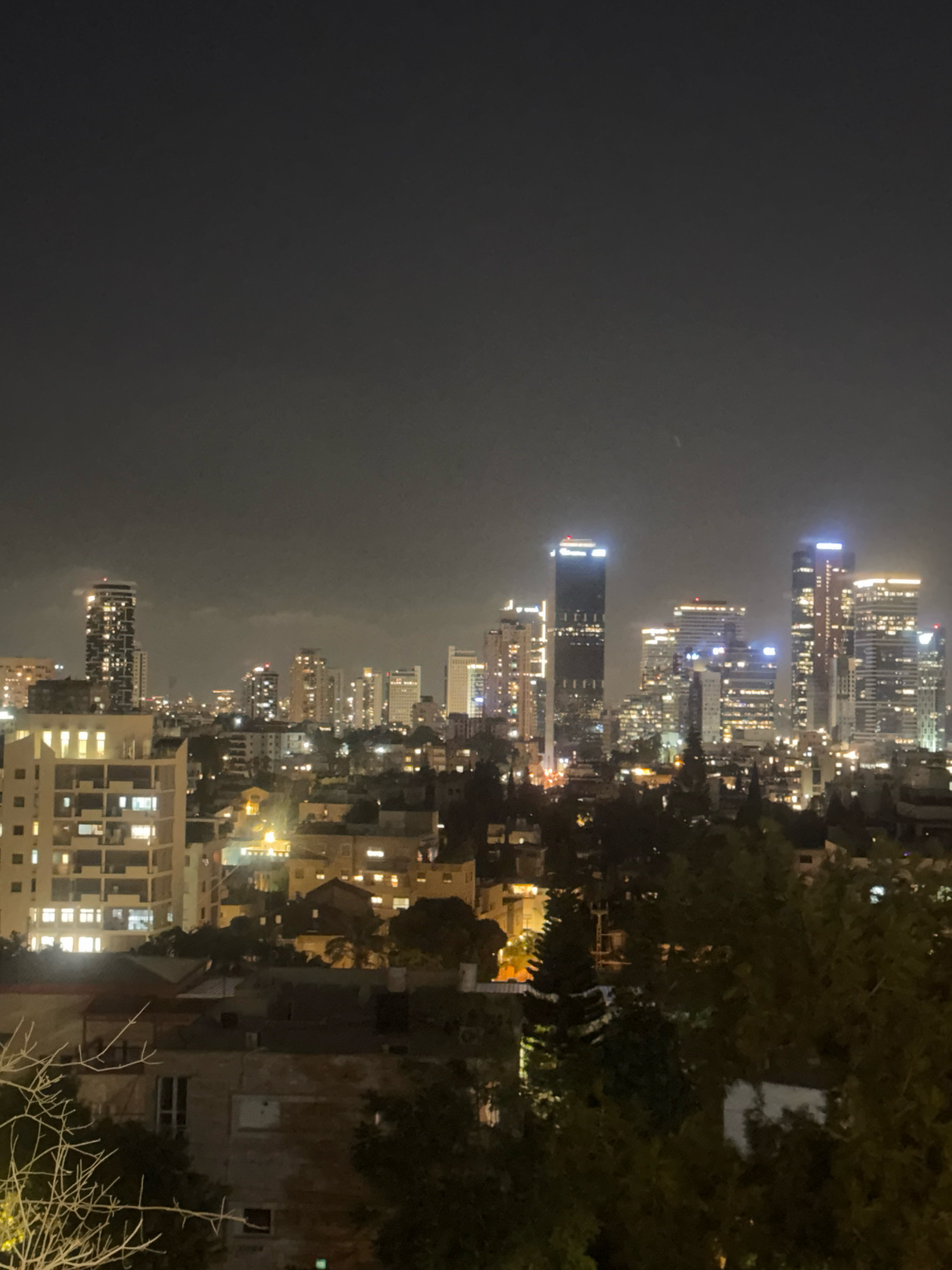 This is a nighttime photograph taken from a high vantage point overlooking the city of Ramat Gan, Israel. The skyline is dominated by numerous illuminated buildings, including several tall skyscrapers. In the foreground, there are dense trees and the tops of lower buildings. The sky is dark and cloudy, with the artificial lights of the city reflecting off the cloud cover. The overall impression is of a sprawling urban landscape at night.