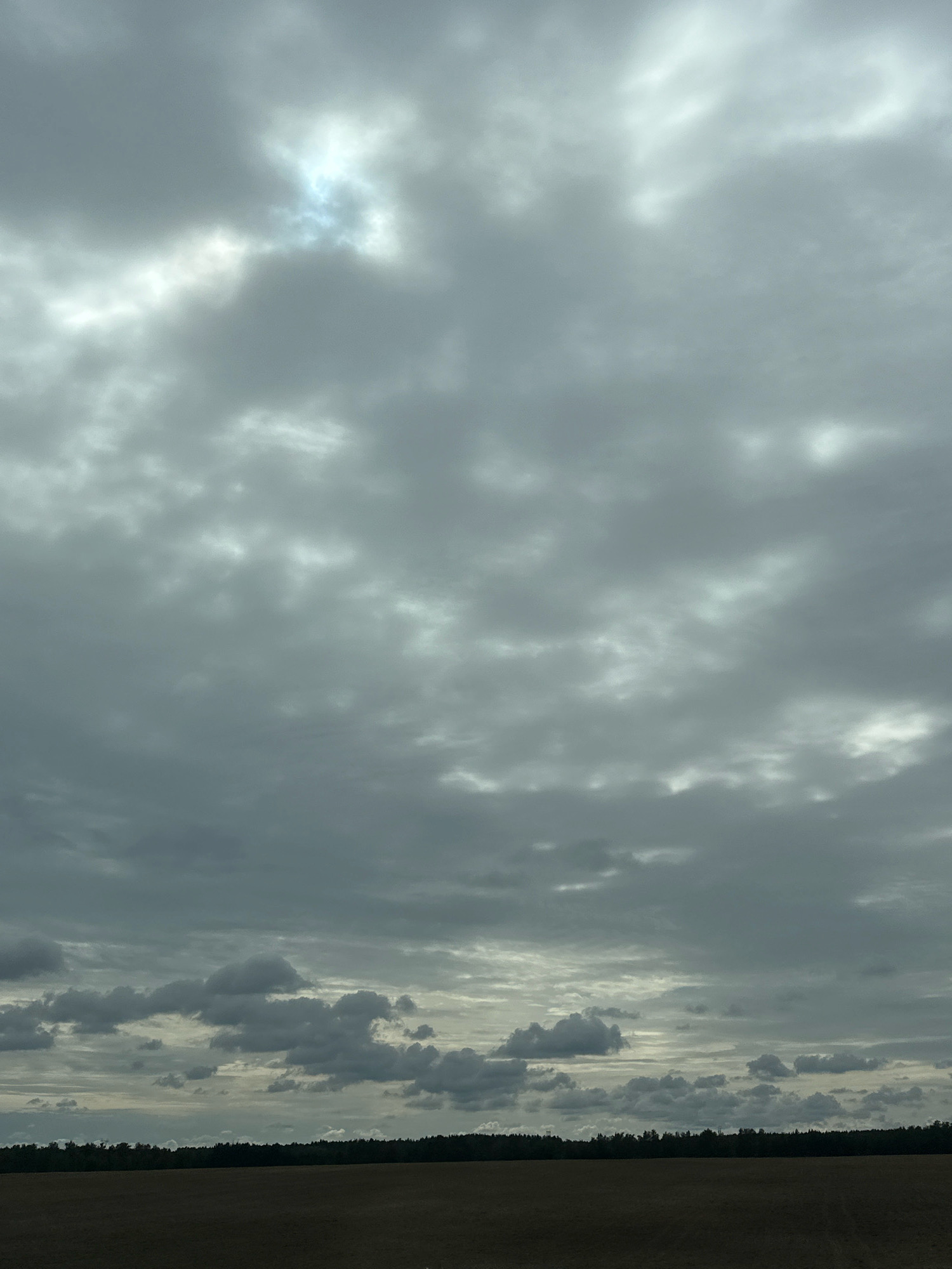 The image depicts a cloudy sky over a rural landscape in Гарбацэвіцкі сельскі Савет, Belarus. The sky is predominantly filled with various shades of grey clouds, with a small patch of brighter, possibly sunlit, sky visible towards the upper left. Below the sky, a dark line of trees forms a horizon, suggesting a forest or dense vegetation. The foreground consists of a vast, dark field, possibly agricultural land, that appears to be barren or harvested. The overall scene conveys a somber or overcast atmosphere. There are no people, animals, or specific structures visible in the image.