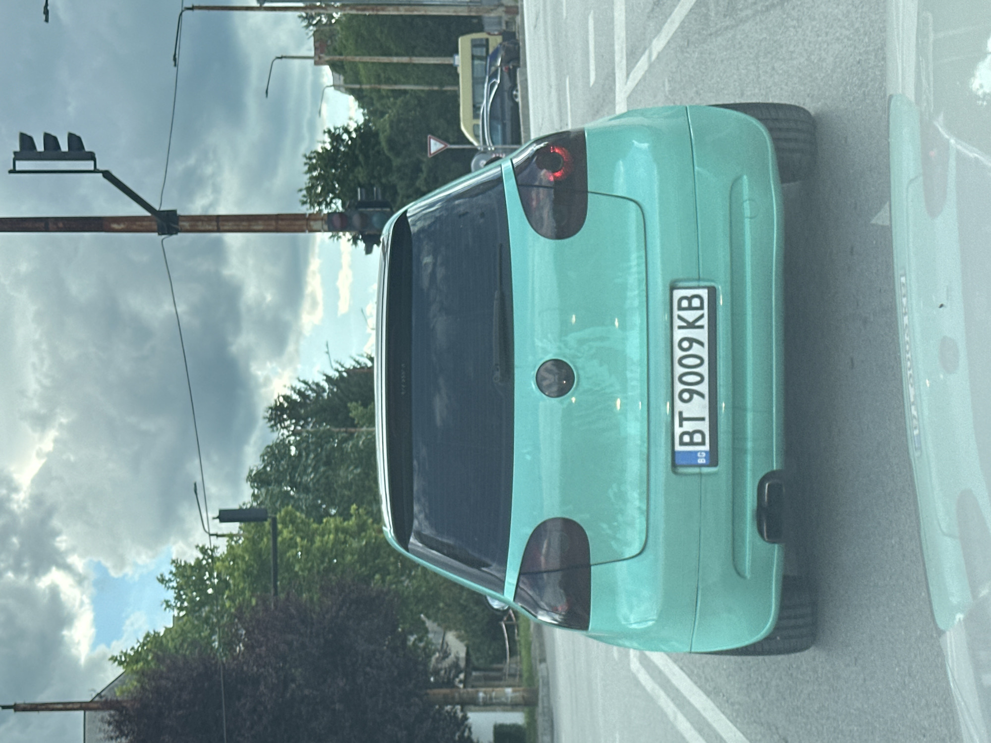 A turquoise Volkswagen Golf is viewed from the rear, stopped at a traffic light in Veliko Tarnovo, Bulgaria. The car has tinted taillights and a license plate that reads "BT 9009 KB". In the background, there are trees, buildings, and traffic lights. The sky is cloudy.