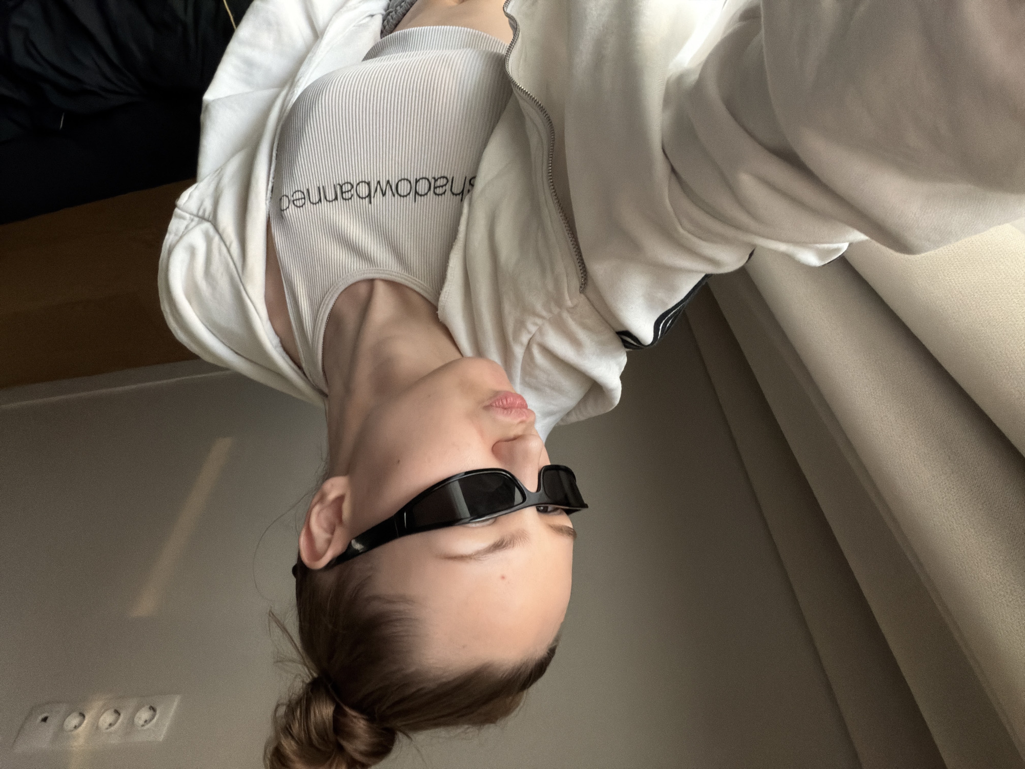 A young woman with her hair in a bun is seen in an upside-down selfie wearing black sunglasses, a white tank top with "shadowbanned" printed on it, and a white zip-up hoodie. The background features a wall with electrical outlets and a curtain. There is no discernible evidence to suggest the location is in the city of Moscow, Russia.
