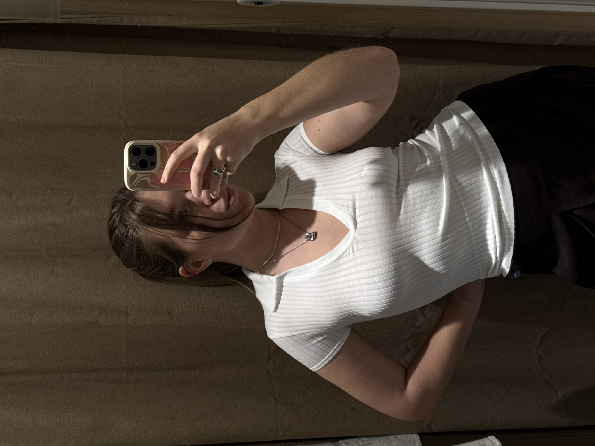 A young woman lies on a tan fabric surface, possibly a bed or couch. She wears a white ribbed top with a collar and short sleeves and a black bottom. She holds a smartphone with a decorative case in her right hand, presumably taking a selfie. A silver necklace adorns her neck. The environment appears to be indoors, with a window or light source casting shadows on the fabric surface. The location in Belgrade, Serbia, cannot be determined from this image alone.