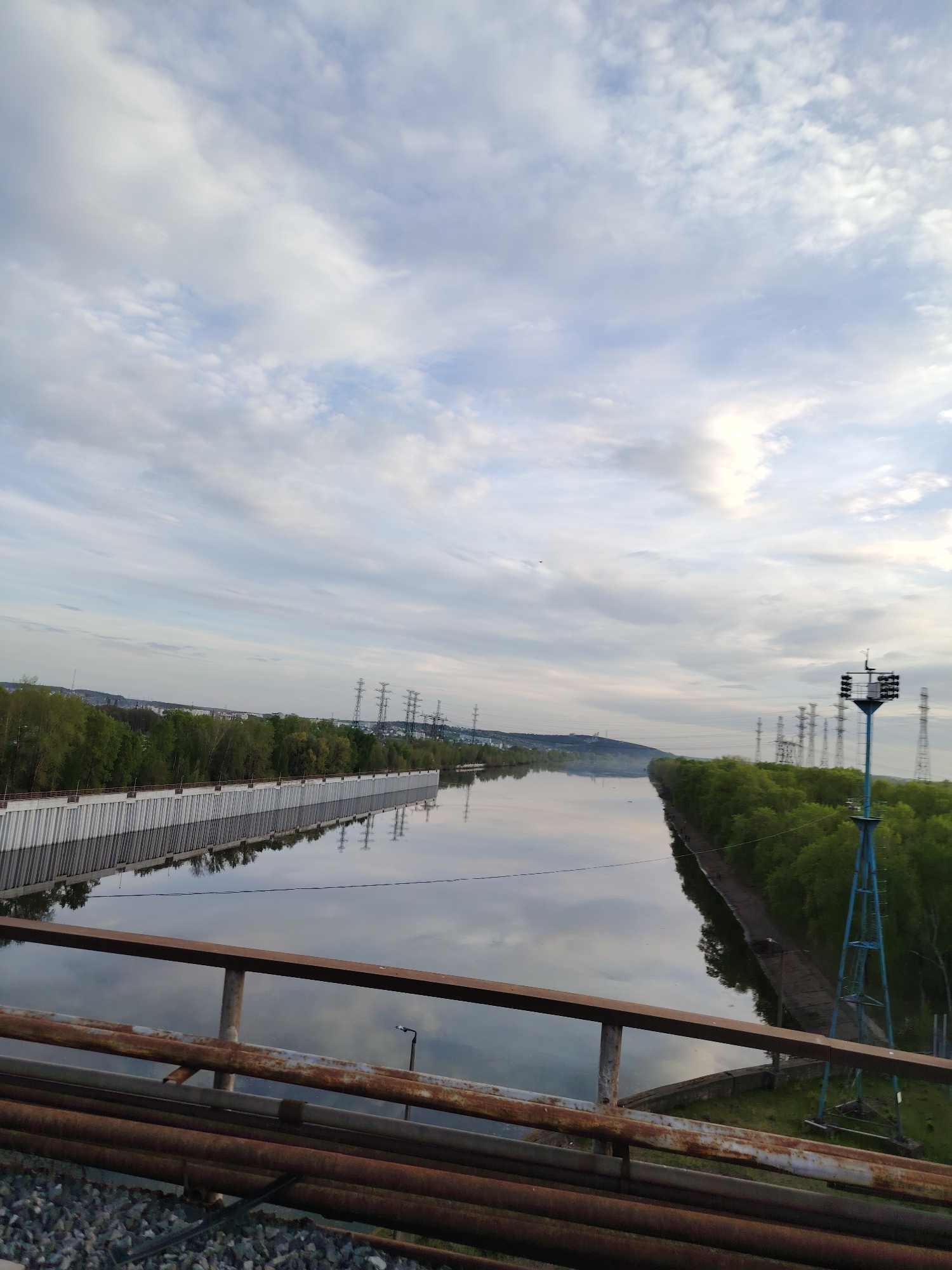 The image shows a wide canal in Chaikovsky, Russia, viewed from an elevated position, likely a bridge. The canal stretches into the distance, reflecting the cloudy sky above. The water is relatively calm.
On the left bank of the canal is a concrete wall structure and beyond it, a row of trees. On the right bank, dense green trees line the water's edge, interspersed with utility poles.
In the foreground, a rusty metal railing and piping obscure the lower portion of the frame. Below the railing, there is a pile of gray gravel. On the right side, a blue, metal tower structure stands.
In the distance, more utility poles are visible on both sides of the canal, with a suggestion of urban development and a low hill in the background. The sky is covered with gray and white clouds.