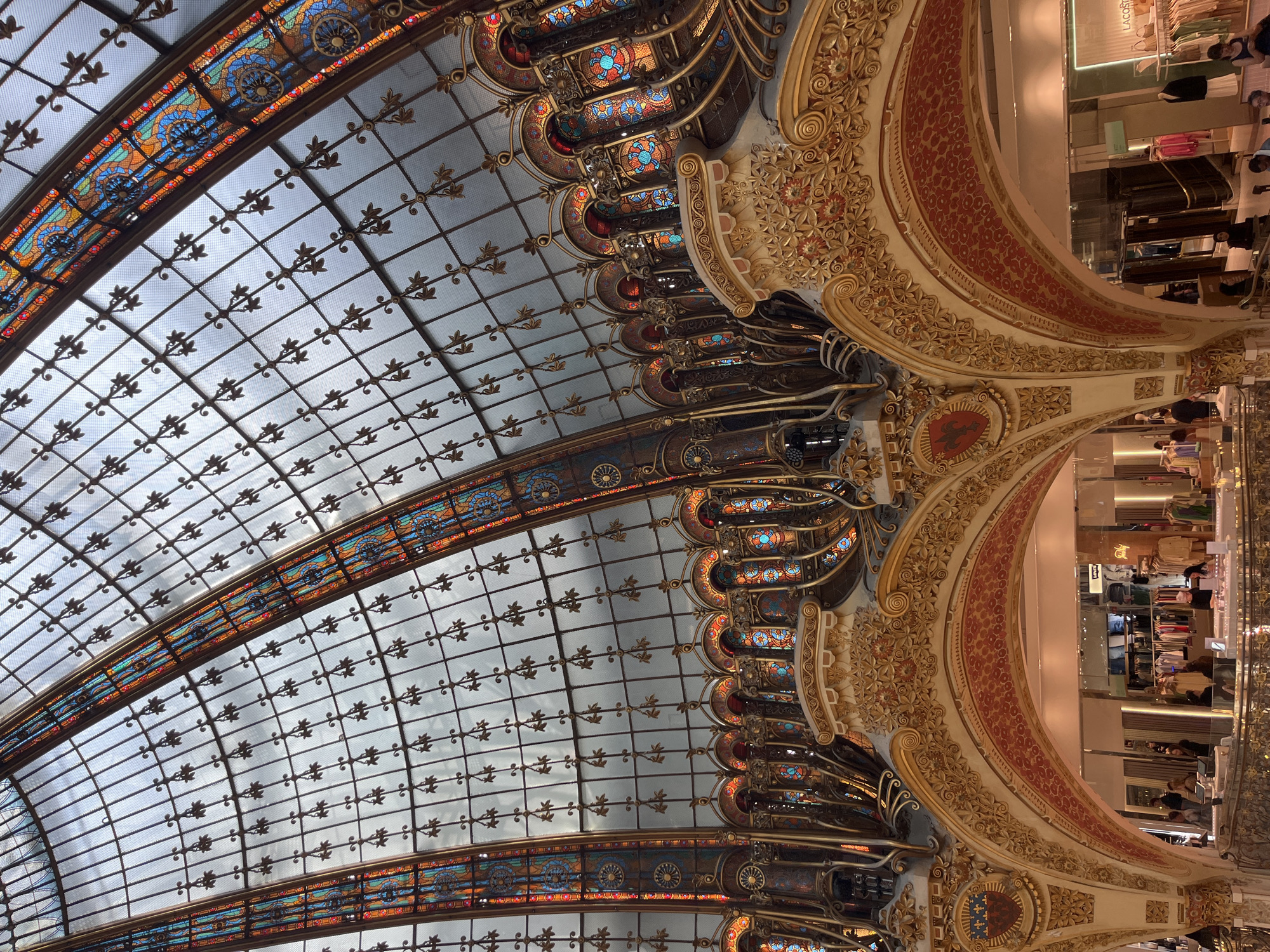Here is a detailed description of the image:
The image captures the interior of a grand, ornate building, likely a department store, in Paris, France. The perspective is from below, looking up at the elaborate ceiling and upper-level architecture.
The ceiling is a massive glass dome, intricately patterned with a grid of metal framework, adorned with decorative leaf-like elements. The glass panels allow diffused light to filter through, giving the scene a soft, ethereal glow. Bands of colorful stained glass run along certain structural lines of the dome, adding visual interest and vibrant hues.
To the right, the architecture of the upper levels is visible. These are characterized by sweeping, curved forms, heavily ornamented with gilded floral motifs and red patterned inserts. The detailing suggests a Beaux-Arts or Art Nouveau style.
In the background, glimpses of merchandise displays and shoppers can be seen, indicating the commercial nature of the space. A sign with the word "LACOSTE" is visible in the upper-right corner.
The overall impression is one of opulence and grandeur.