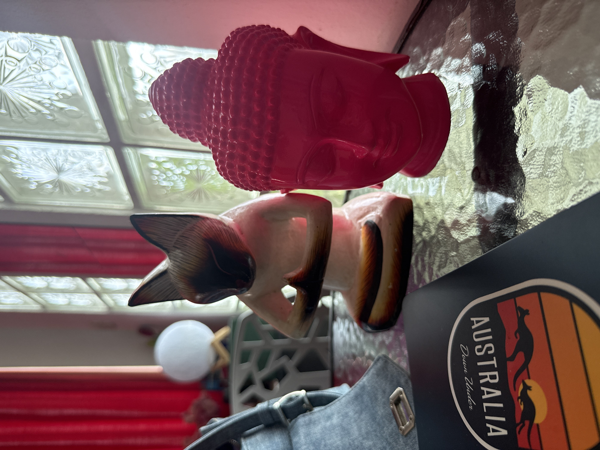 On a textured glass table, an indoor still life scene, likely from Baan Chaweng Noi, Thailand, unfolds under bright daylight. Prominently positioned on the left side of the frame is a vibrant red statue of a Buddha head, distinguished by its serene expression and tightly curled hair pattern (ushnisha).
Adjacent to the Buddha head and slightly to its right, a wooden statue of a reclining cat is visible, possibly a Siamese cat, characterized by its lighter body and contrasting darker markings on its ears, paws, and tail. In the mid-ground on the right, a black rectangular object displays a 'AUSTRALIA Down Under' sticker, which features silhouettes of two kangaroos against a graphic sunset of orange and yellow stripes.
Portions of a gray bag with a buckle and a metallic loop are visible in the bottom right corner of the frame. The background includes a section of patterned glass blocks, likely part of a window or wall, which refract and reflect light. Blurred red horizontal elements, possibly blinds or a piece of furniture, are seen in the lower left background, along with a soft white spherical object that could be a light fixture. No specific activity is taking place; the image captures an arrangement of personal and decorative objects.