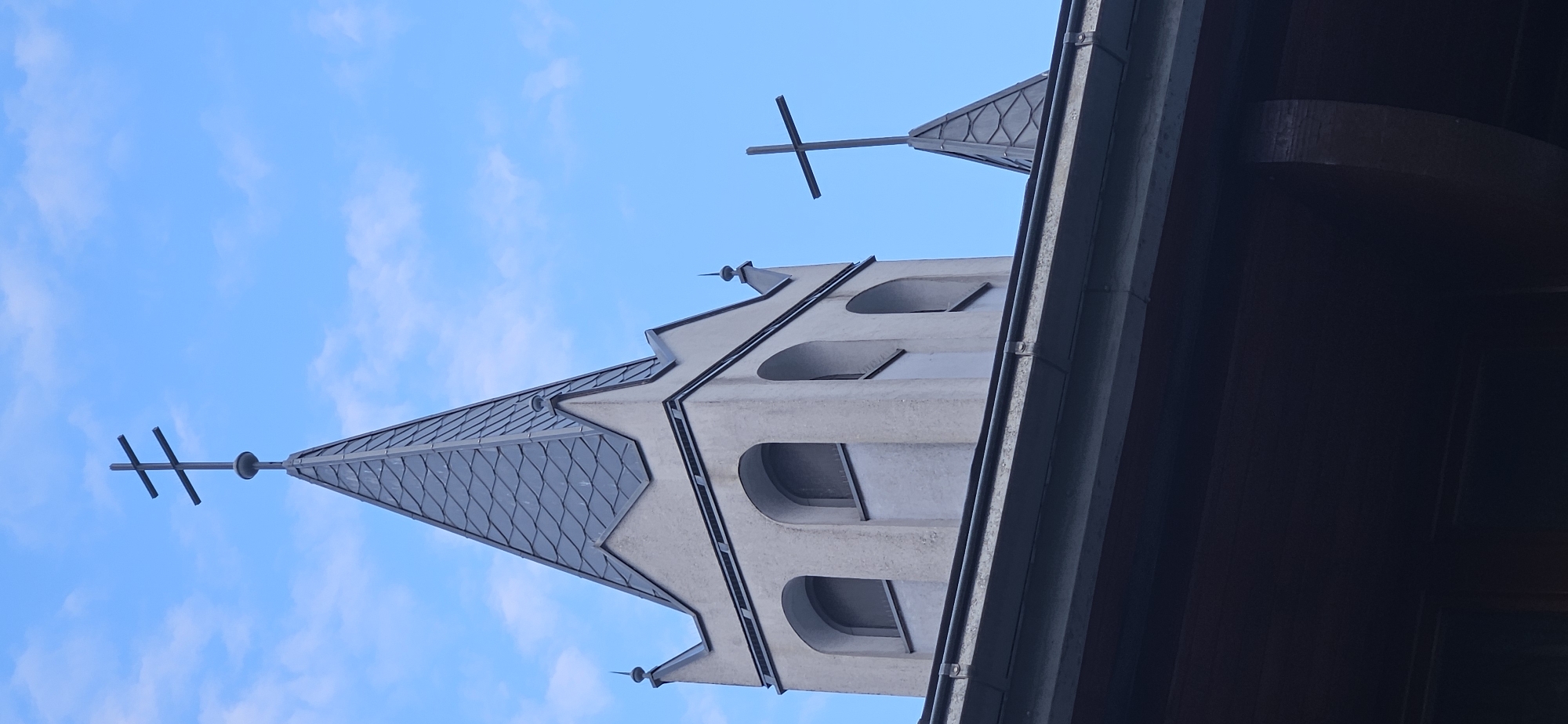 The image is a low-angle, vertical shot of a church tower against a bright blue sky with scattered clouds. The tower is made of light-colored stone and features a steep, grey, slate-tiled roof that tapers to a point. A double-barred cross, also known as a patriarchal cross, sits atop the main spire. Below the spire, the tower has arched windows. To the right of the main tower, a smaller, similarly shaped spire with a single-barred cross is visible. The foreground is dominated by the dark, sloped roof of a building, possibly part of the church itself or an adjacent structure, with a gutter running along its edge. The overall impression is of a clear day in a city with traditional religious architecture. The architecture of the church tower and the presence of crosses suggest it is likely a Christian church. This imagery is consistent with religious buildings found in Hévíz, Hungary, a city known for its spa and the Saint Andrew's Church.