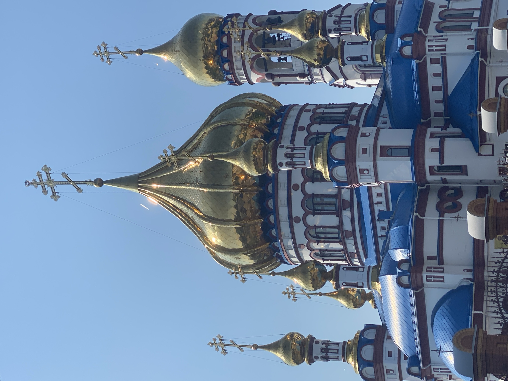 The image shows a close-up, low-angle shot of the Church of the Transfiguration in Novokuznetsk, Russia. The church has multiple golden domes with crosses, white walls, and blue roofs. The sky is clear and blue. The church is adorned with red trim and decorative elements. In the foreground, parts of a dark metal fence and brick pillars are visible.