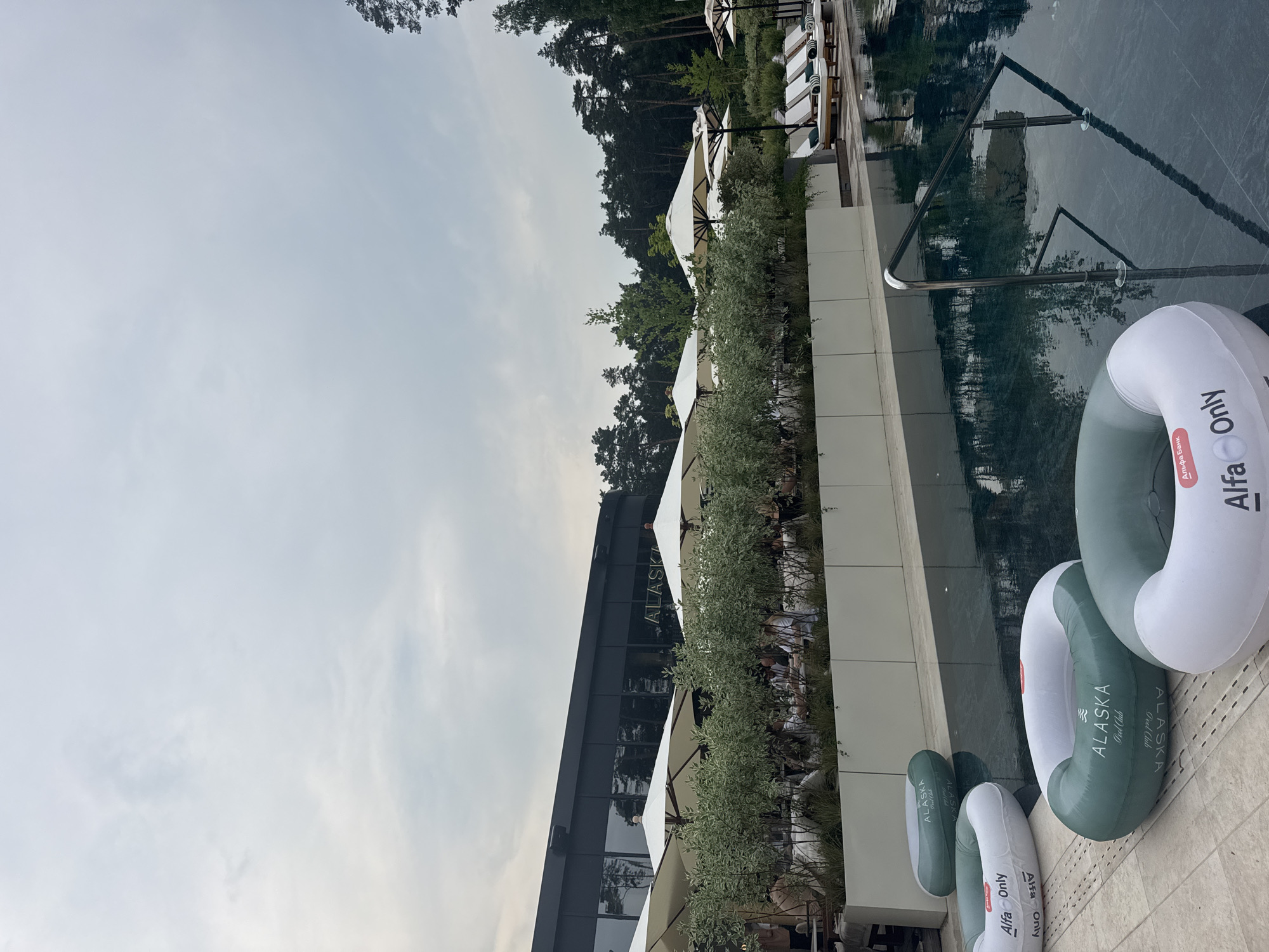 In Razdory, Russia, at a poolside establishment, the scene is set under an overcast sky. The foreground features a swimming pool with three inflatable rings partially submerged in the water, two of them displaying the "Alfa Only" logo and one with "ALASKA Pool Club". The pool's edge is lined with beige tiles. To the left of the pool, on the tiled surface, lie two more inflatable rings, one white with green lettering "ALFA Only" and another white and green with "ALASKA Pool Club" branding. Beyond the pool, a white planter box contains dense green shrubbery, separating the pool area from a seating area. Several white umbrellas are open over tables where people are seated, suggesting an outdoor dining or lounge space. In the background, a modern building with a dark facade and reflective windows is visible, bearing the "ALASKA" logo. Further back, a dense forest of pine trees forms the horizon. Beach chairs are arranged in neat rows to the right of the seating area, indicating a resort or leisure facility. A silver handrail provides access to the pool.