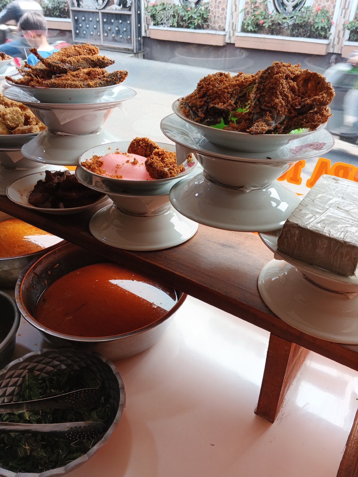 The image depicts a display of food items on a wooden shelf in what appears to be a food establishment. In the foreground, there are several bowls and containers filled with food. One large metal bowl contains a reddish-orange liquid, likely a soup or sauce. Next to it, a smaller decorative metal bowl holds green leafy vegetables, with metal tongs resting inside. To the right, a rectangular object wrapped in plastic wrap sits on a white plate.
Above these, stacked white plates and bowls hold various food items. One bowl contains a pinkish substance with brown crumbs sprinkled on top, possibly a dessert. Another bowl holds dark, cooked meat. Further up, a stack of white bowls displays fried fish, some covered in a brown, crispy coating. The food is arranged in a tiered display.
In the background, through a window, a person wearing a blue shirt is visible, facing away from the camera. The setting appears to be indoors with natural light coming from the window. The overall impression is a visually rich display of Indonesian cuisine, likely in a restaurant or food stall. While the image provides a detailed look at the food and its presentation, it does not contain explicit elements to pinpoint the specific location within Gandasari, Indonesia.