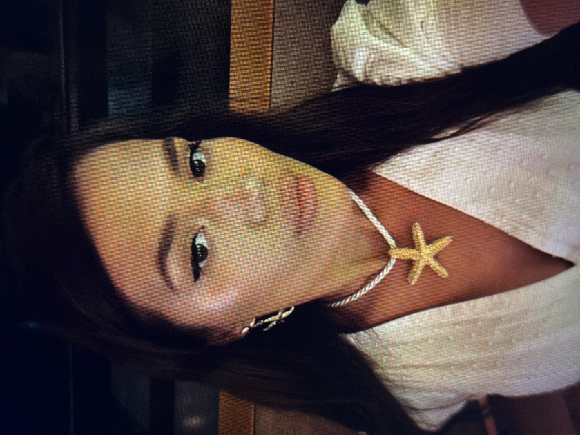 A medium shot captures a woman from the chest up, facing the camera. She has long, dark hair and is wearing a white textured top with puffed sleeves. A necklace with a gold starfish pendant rests on her chest, and she has hoop earrings. Her makeup includes winged eyeliner and lip gloss. The background is dark and out of focus, with hints of orange and brown tones, possibly indicating an indoor setting with decorative elements. There is no identifiable information about the location in Sirius, Russia.