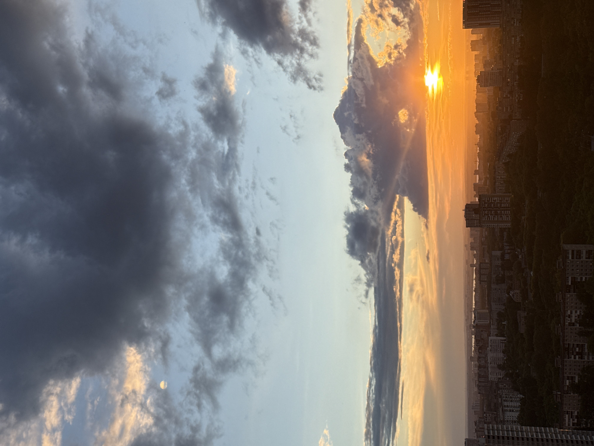 This image depicts an aerial view of a city skyline at sunset, featuring a prominent sun partially obscured by dramatic clouds. The sky transitions from a pale blue at the top to vibrant oranges and yellows near the horizon. Dark, heavy clouds dominate the upper portion of the sky, with lighter, more wispy clouds scattered throughout. Below the sky, a dense urban landscape is visible, characterized by numerous buildings of varying heights, including apartment blocks and taller modern structures. A band of lush green trees separates the lower buildings from the distant horizon. The scene is illuminated by the warm glow of the setting sun, casting long shadows and highlighting the urban environment. The city of Perm, Russia, is known for its industrial and residential areas, and this image captures a typical view of its developed landscape during a picturesque sunset. There are no people or animals visible in the image.