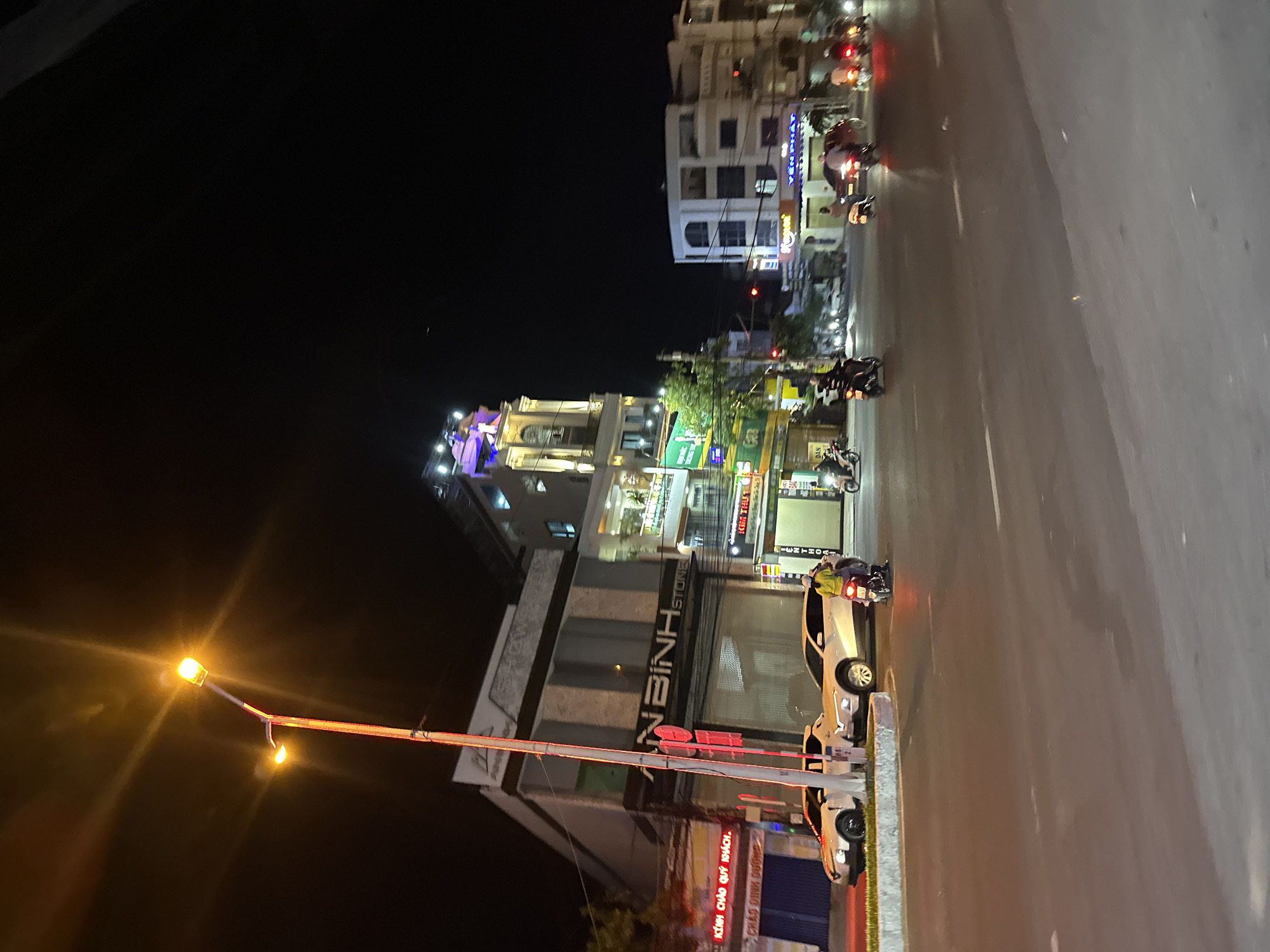 A nighttime street scene in Bac Nha Trang Ward, Vietnam, shows a view along a road with buildings and motorbikes. Buildings line the street, illuminated by lights, with a business called "AN BINH STONE" visible. Cars are parked along the road, and motorbikes are in motion. A traffic light stands near the buildings, and overhead streetlights brighten the scene. Signage is present, written in Vietnamese. The general atmosphere is urban and illuminated by artificial light at night.