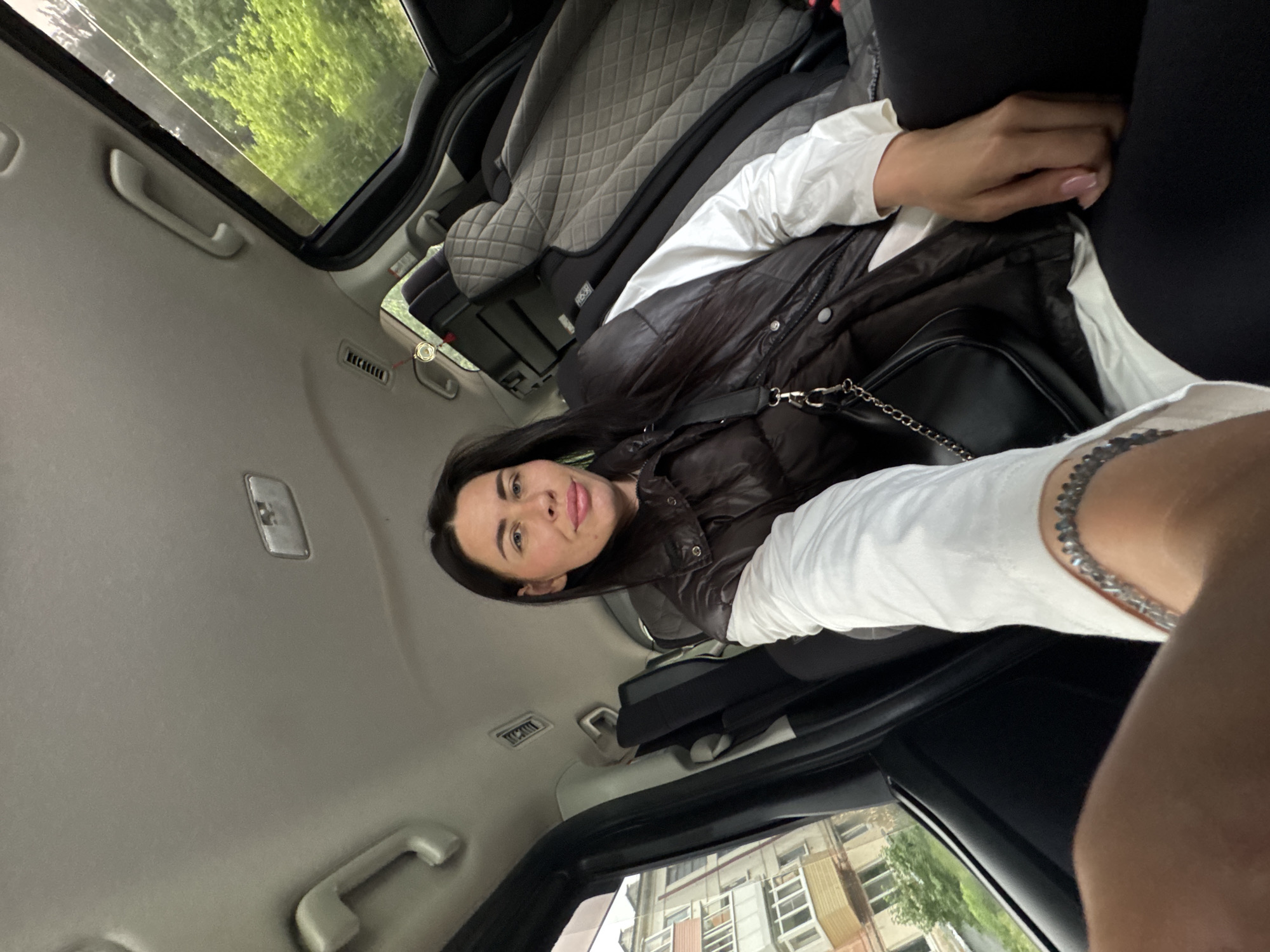 A woman is seated in the back of a car, looking towards the camera. She has long dark hair and is wearing a brown puffer vest over a white long-sleeved shirt and black leggings. A black purse with a chain strap is slung over her shoulder. Her left hand rests on her thigh, and a silver bracelet is visible on her left wrist. The interior of the car shows quilted grey seat covers and dark grey interior panels. Outside the car window, buildings and trees are visible, suggesting an urban or suburban setting. The image appears to have been taken during the daytime.