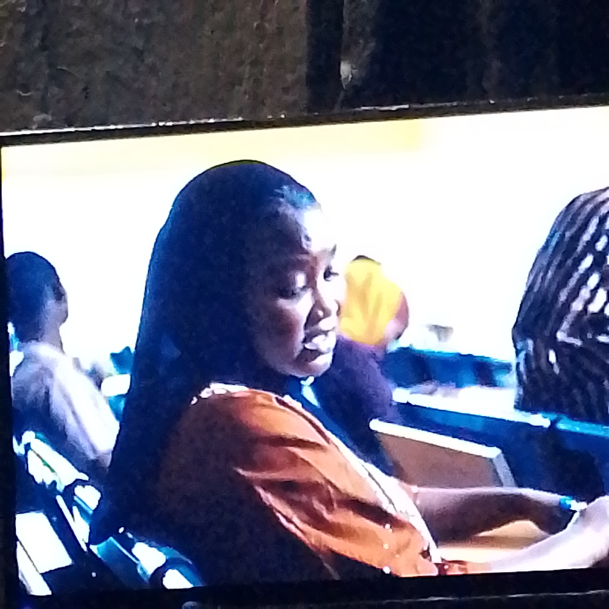 A woman is sitting in what appears to be a row of seats, possibly at a public event or gathering. She has dark hair and is wearing an orange top with small white details. Other individuals are visible in the background, including one with a striped shirt and another with a yellow object behind them. The scene is indoors, likely in Unknown, Unknown.