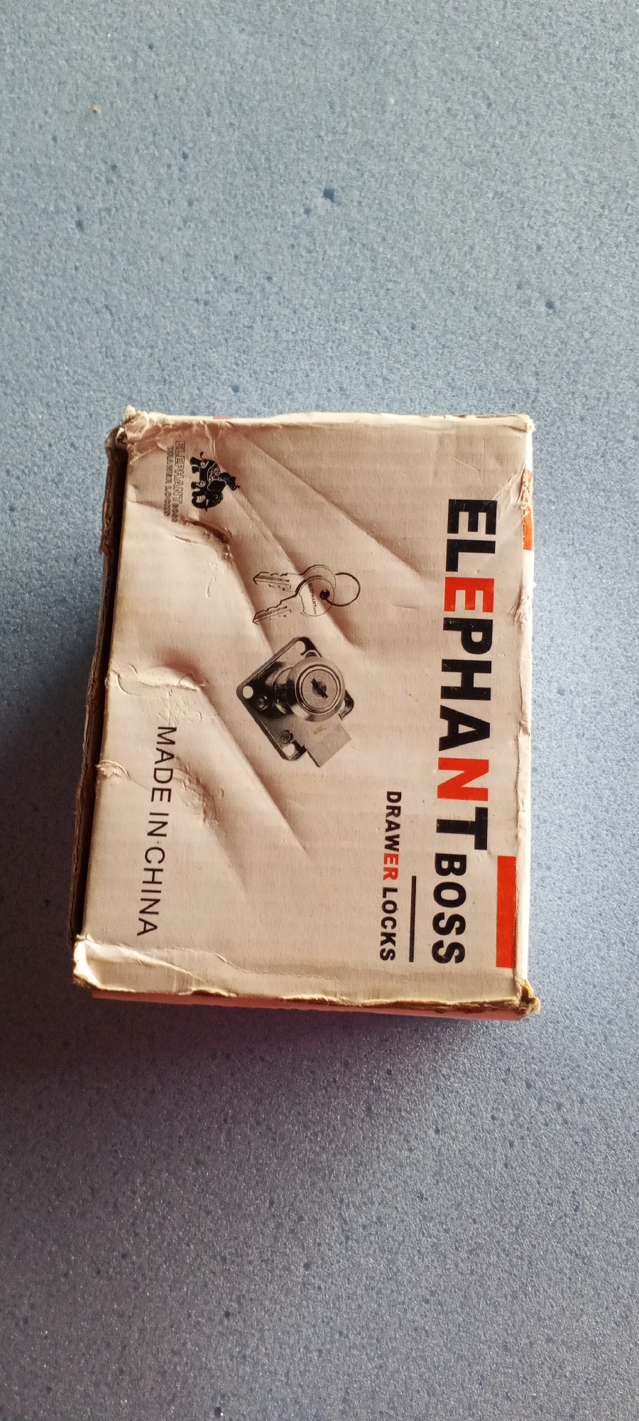 This media file shows a close-up, top-down view of a damaged cardboard box with text and images printed on it, resting on a textured surface. The box appears to contain drawer locks, as indicated by the text and a printed illustration of a lock and keys. The box has significant wear and tear, with crumpled edges and creases.
Visible text on the box includes "ELEPHANT BOSS" printed vertically in large black and red letters, and "DRAWER LOCKS" printed horizontally in smaller red letters. There is also text that reads "MADE IN CHINA" printed vertically on the left side of the box. An illustration of an elephant is also visible in black.
The background is a uniform, lightly textured surface, possibly a table or floor, with a bluish-grey hue. The lighting is even, suggesting it is likely indoors or under diffused daylight. No people are visible in the image. The overall impression is that of a product package that has experienced some rough handling.