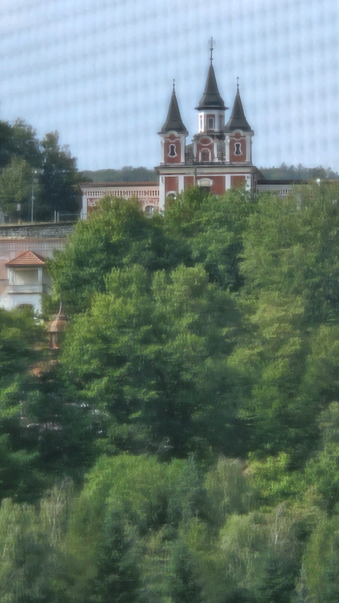 The image shows a view of the Saint Nicholas Greek Catholic Cathedral in Prešov, Slovakia. The cathedral is a prominent religious building with a distinctive architectural style featuring multiple spires and red and white facades. It is situated on a hill, surrounded by lush green trees. The foreground of the image is dominated by dense foliage, with hints of a lower building or structure partially visible to the left. The sky is overcast with a pale blue hue. The overall scene depicts a tranquil, architectural, and natural landscape.