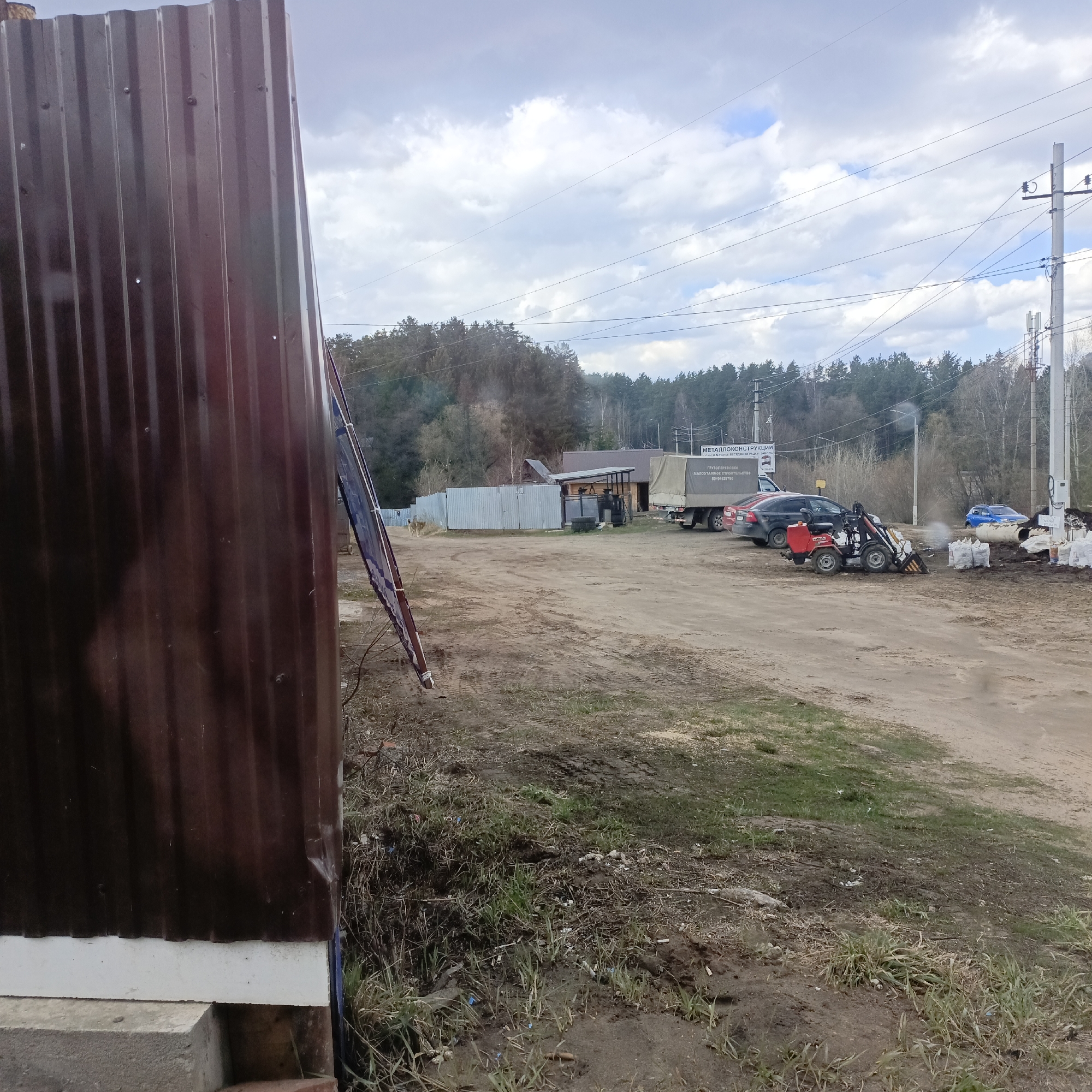The image depicts an outdoor scene in an unidentified urban area. In the foreground, there's a corrugated metal structure, possibly a shed or building extension. The ground around the structure is a mix of dirt, grass, and some scattered debris.
Moving into the midground, there's a dirt road or yard area. Several vehicles are present, including a truck with a covered bed, a red car, a black car, a blue car, and a small red front-end loader or forklift. There are also piles of white bags, possibly containing materials like soil or fertilizer. Smoke or dust is seen near the front-end loader, suggesting recent activity.
In the background, there's a fence, some buildings, and a wooded area with a mix of evergreen and deciduous trees. Overhead, there are power lines and utility poles extending across the scene. The sky is cloudy, indicating an overcast or partly cloudy day.