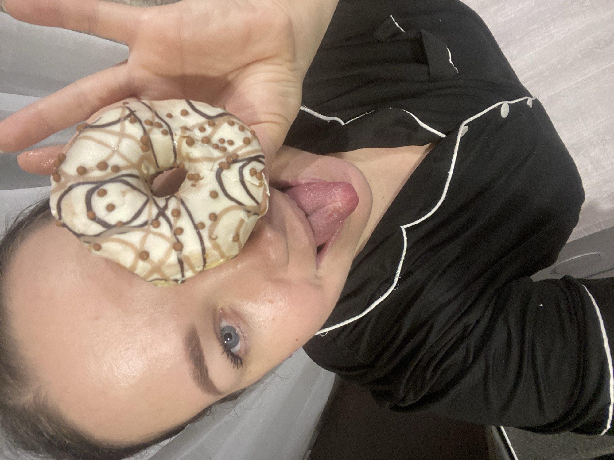 A female subject is depicted in a close-up, upward-angled selfie, holding a donut directly in front of her face, partially obscuring her left eye and forehead. Her visible right eye is blue. She is sticking out her tongue. The donut features white icing with irregular drizzles of light brown and dark brown frosting, and is covered with small, spherical brown sprinkles. The subject is wearing a black garment, likely pajamas, with white piping along the collar and button placket. Her right hand, holding the donut, is partially visible. The background consists of indistinct indoor elements, including a light-colored surface on the left and a darker area on the right, suggesting a casual, personal setting. The precise location within the city of Sochi, Russia, cannot be determined from the visual information present in the image.