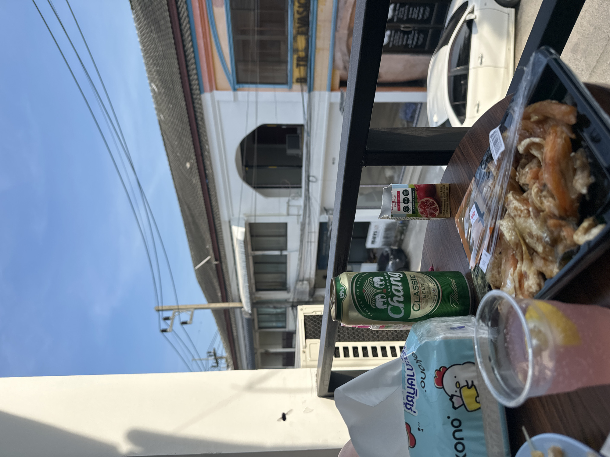 The image shows a table setting with food and drinks in what appears to be Baan Chaweng, Thailand. On the table, there's a plastic container of what looks like fried food, a can of Chang Classic Beer, a carton of Tipco fruit juice, a plastic cup with a pink liquid and a lemon slice, and a box of Kono tissues. The backdrop shows a white building with an arched doorway, power lines, and a blue sky. A white car is partially visible behind the table. The scene suggests an outdoor eating area or balcony with a view of the street.