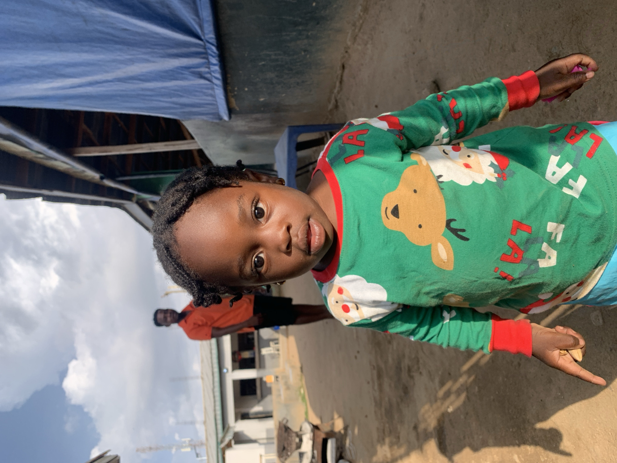 The image shows a young child, likely a girl, of African descent, standing outdoors and looking directly at the camera. She has dark, braided hair and a neutral, observant expression. She is wearing a green long-sleeved shirt with red cuffs, patterned with Christmas motifs including a brown reindeer, Santa Claus faces, and white text that clearly displays "LAFA" on the lower right side. A glimpse of blue clothing, possibly pants, is visible on her right side. In her right hand, she holds a small, light-colored object, possibly a seed or a small stone.
In the background, an adult male, out of focus, is visible standing. He wears an orange shirt and dark shorts. The setting appears to be an outdoor area with a light brown, sandy or dirt ground. Above and to the child's left, a blue tarp structure is visible. Further in the background, out-of-focus buildings and a bright blue sky with scattered white clouds indicate clear, sunny daytime weather. The scene suggests a community or residential environment.