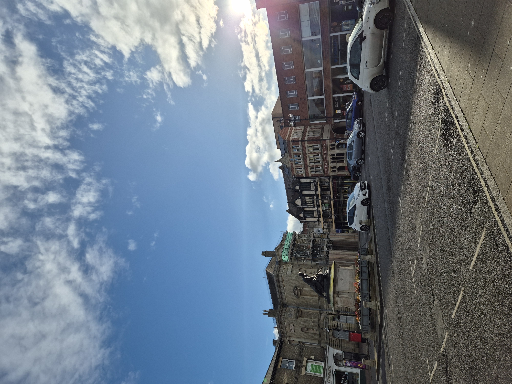 The image depicts a street scene in West Suffolk, United Kingdom, under a bright, partly cloudy sky. The road is lined with parked cars on one side and various buildings on the other. A historical stone building, possibly a monument or civic structure, stands prominently along the street, partially obscured by scaffolding. Adjacent buildings include shops, and a taller red-brick structure with multiple stories. The scene is bathed in sunlight, casting shadows along the road and highlighting the textures of the building facades.