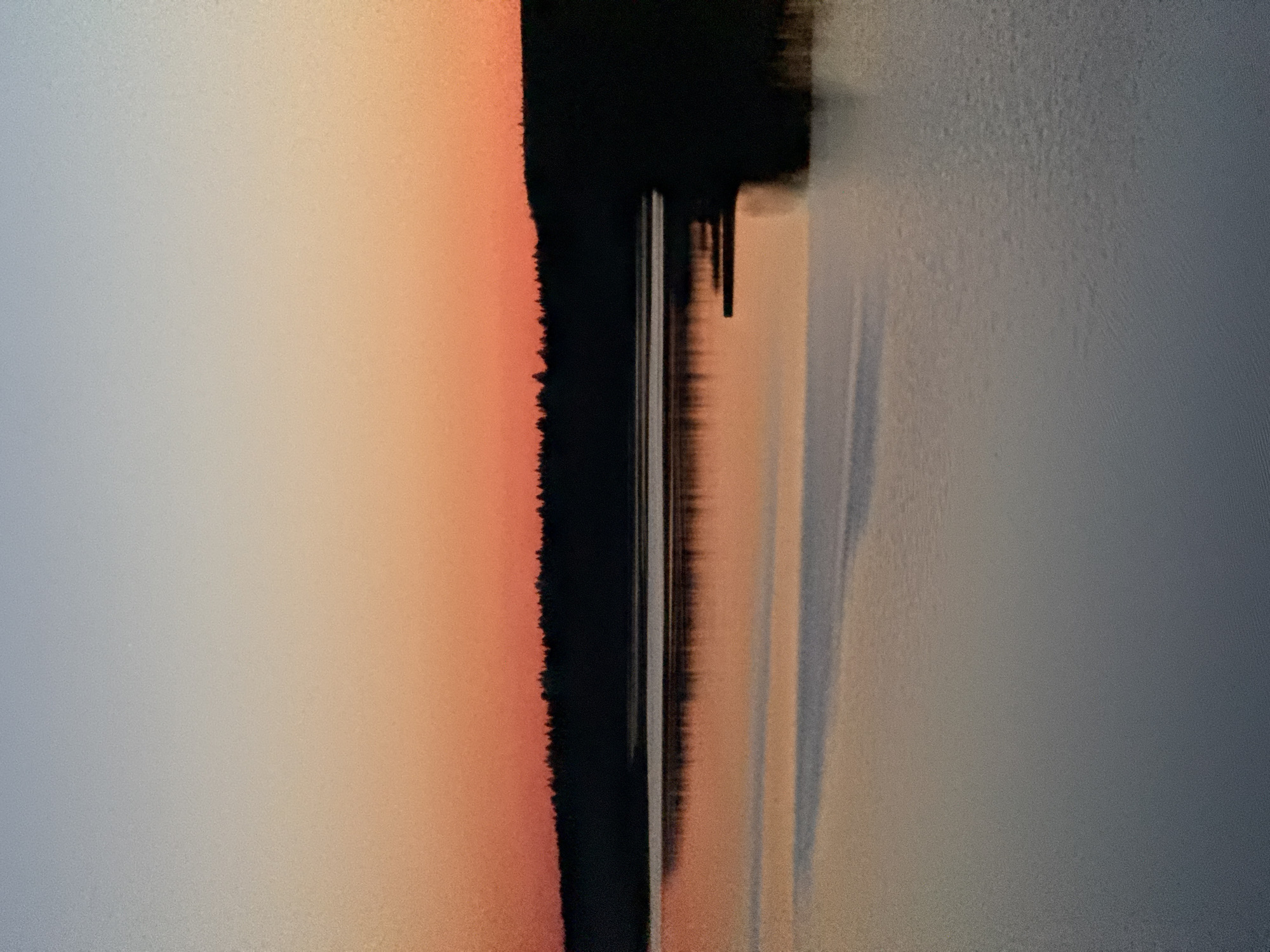 The image shows an abstract composition with a focus on color and texture. On the left, there is a soft gradient transitioning from light grey to a muted orange hue. A thick, irregular black stripe runs vertically, dividing the left and right portions of the image. To the right of the black stripe, the image transitions into a textured grey area, with elongated, blurred lines and shadow-like effects extending downwards. There is no indication of the city of Votkinsk or any people present in the image. The scene appears to be a close-up, possibly of an artwork or surface with distinct textural and color elements.