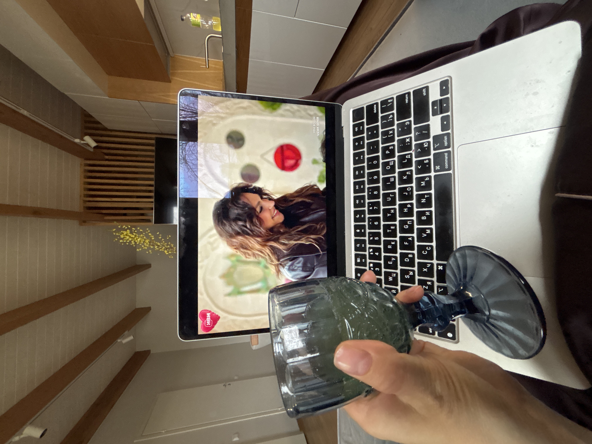 The image presents a first-person view of an indoor setting, focusing on a person's hand holding a dark blue stemmed glass. A silver MacBook laptop is open in front of the person, displaying a video on its screen. The video shows a woman with long, wavy brown hair, smiling and looking to the left. Visible text on the screen includes "ИВИ" (IVI), a Russian online video service logo, and "YENİ BÖLÜM UZAK ŞEHİR," which translates from Turkish to "NEW EPISODE FAR AWAY CITY."
The laptop's keyboard features both Latin and Cyrillic characters, indicating a Russian keyboard layout, consistent with the given location context of Chaikovsky, Russia. In the background, a modern interior space is visible, characterized by light-colored walls, horizontal wooden shelves, and what appears to be a kitchen counter with a stainless steel faucet and a partially visible "BON AQUA" water bottle. A small bouquet of yellow flowers, possibly mimosa, rests on one of the shelves. The scene conveys a sense of relaxed entertainment in a home environment.