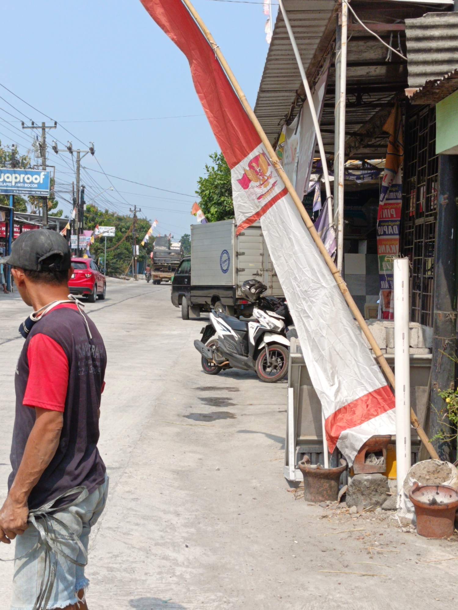 A man wearing a black cap, red shirt, and blue shorts stands in the foreground, facing away from the camera and looking down the street. He appears to be holding a bundle of straps. Behind him, a large red and white flag hangs from a bamboo pole. The street is paved with concrete and lined with buildings and utility poles. A red car is parked on the left side of the street, and a black truck with a white box is visible further down. Several motorcycles are parked near the buildings on the right. The scene appears to be in a commercial or residential area of Manisrenggo, Indonesia, likely during the daytime, as indicated by the bright sunlight and shadows.
