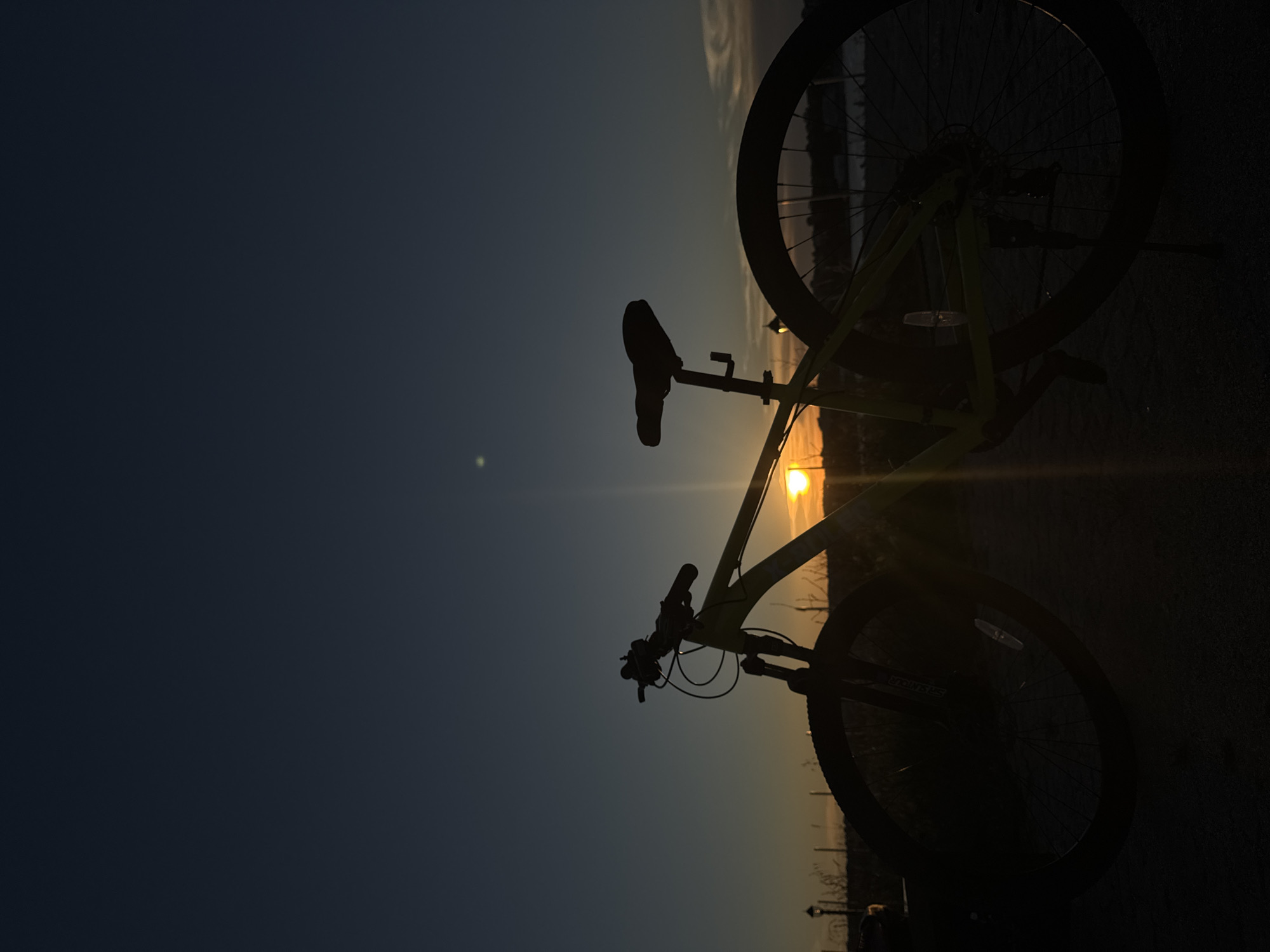 A silhouette of a yellow mountain bike is captured at sunset against a dark blue sky. The sun, a bright orange disc, is positioned just above the horizon, casting a warm glow and creating lens flare. The bike is parked on a paved surface, with hints of grass and a distant body of water visible. The overall scene is serene, with the low sun suggesting the end of the day. The location is identified as Tsarevo, Bulgaria.