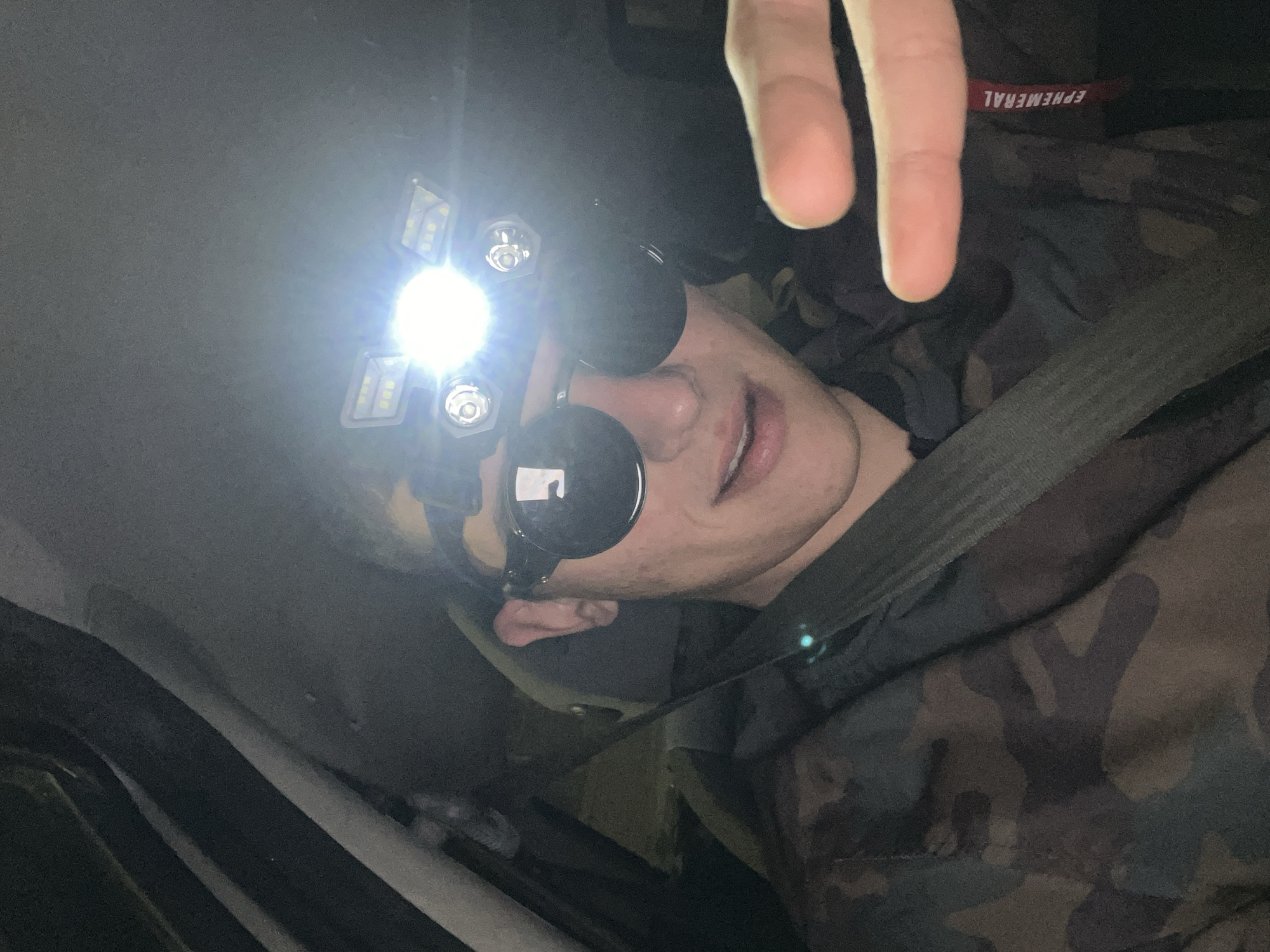 A person wearing a camouflage jacket, sunglasses, and a headlamp is sitting in what appears to be a car. The headlamp is emitting a bright light. A seatbelt is visible across the person's chest. A hand with two fingers extended is also in the frame. The location is not determinable as the image does not contain landmarks or other location information.