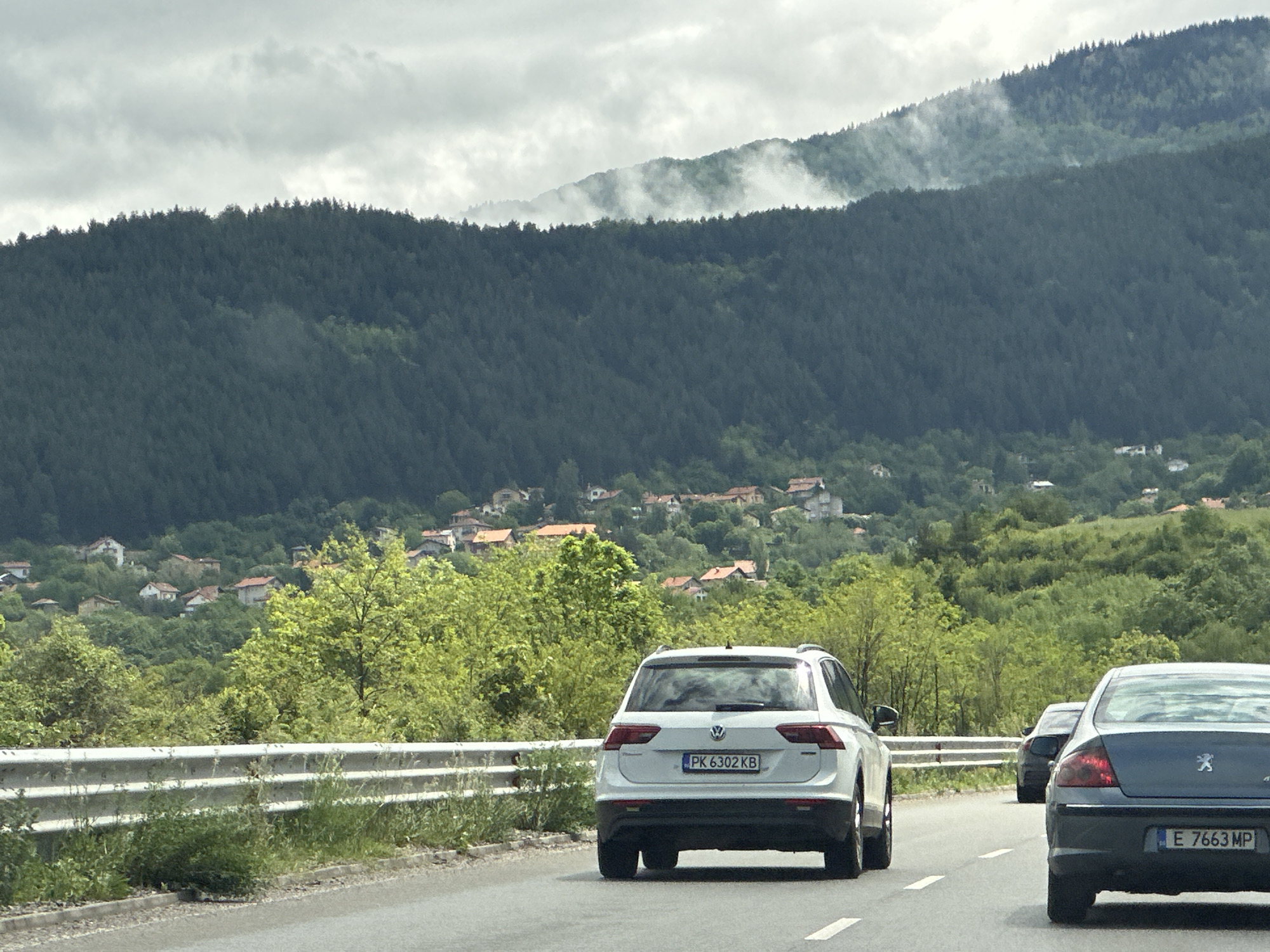 A daytime photograph captures a scenic mountainous landscape with a road in the foreground, likely in Marchaevo, Bulgaria. The sky is overcast with gray clouds.
In the foreground, a two-lane asphalt road stretches into the distance, with a metal guardrail lining its left edge. Three vehicles are visible from behind, driving on the road. A white Volkswagen SUV (likely a Tiguan) occupies the left lane, its license plate reading "PK 6302 KB." Behind it and further to the right, a dark blue or grey Peugeot sedan is in the right lane, displaying the license plate "E 7663 MP." A third, darker vehicle is partially visible further down the road behind the Volkswagen.
Beyond the road, the terrain rises steeply into densely forested mountains, primarily covered with dark green coniferous trees. Lower down, patches of brighter green deciduous trees are also visible, particularly framing a village nestled on the hillsides. The village features numerous houses with reddish-orange tiled roofs, scattered among the trees. Mist or low clouds cling to the upper slopes and ridges of the more distant mountains, adding to the atmospheric quality of the scene. The overall impression is one of a journey through a lush, verdant, and slightly misty mountain region.