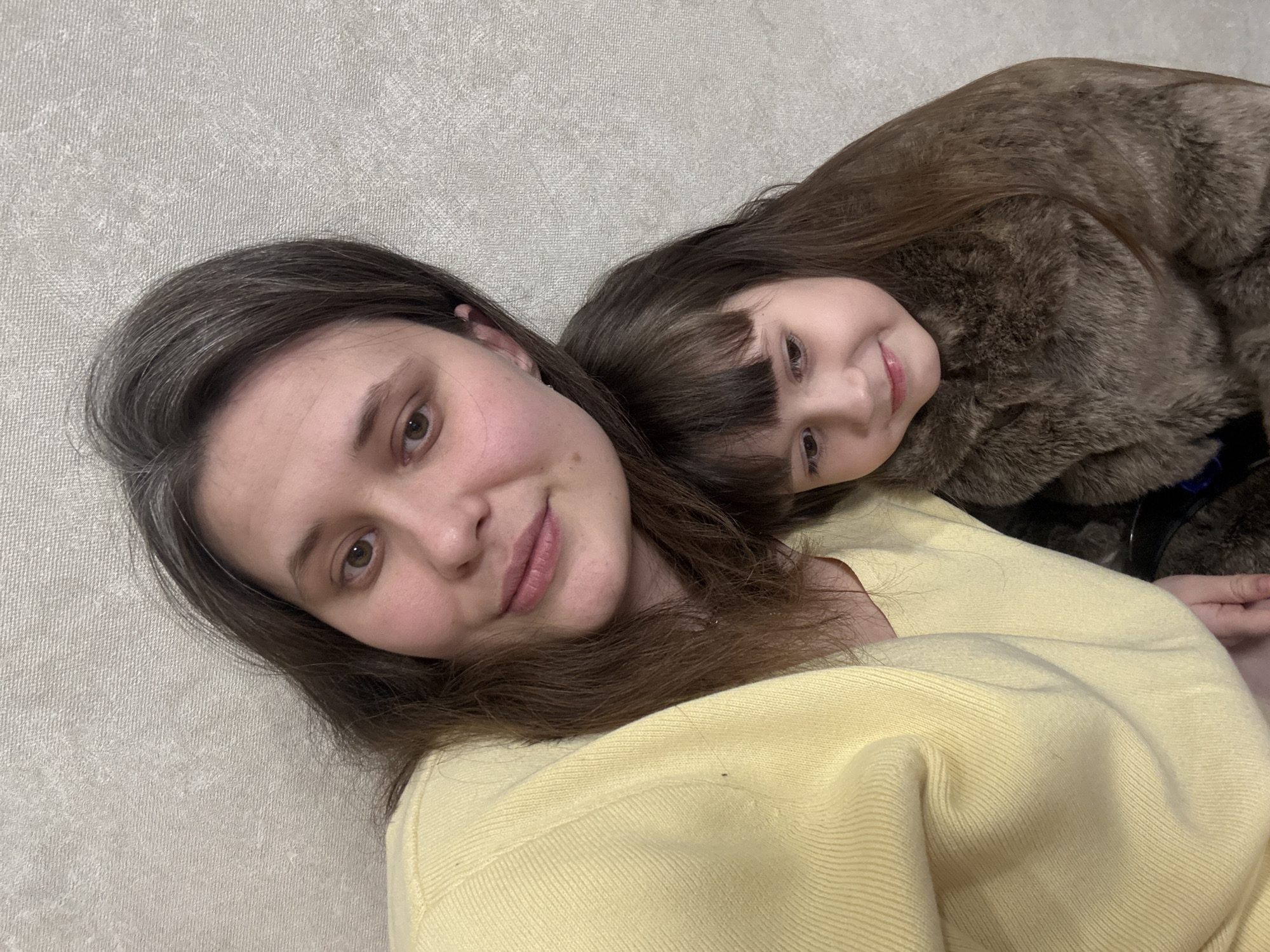 The image displays two individuals, an adult female and a young female child, positioned in close proximity within an indoor setting. The adult female, located on the left side of the frame, has medium-length brown hair with visible grey strands and is wearing a light yellow knitted sweater. She is looking directly at the camera with a slight smile. To her right, the young female child is resting her head on the adult's shoulder or upper torso. The child has brown hair with bangs and is wearing a brown, possibly faux fur, garment. She is also looking towards the camera with a smile. Both individuals have fair complexions. The background consists of a plain, light grey textured wall. The specific location within the city of Chaikovsky, Russia, is not identifiable from the visual content.