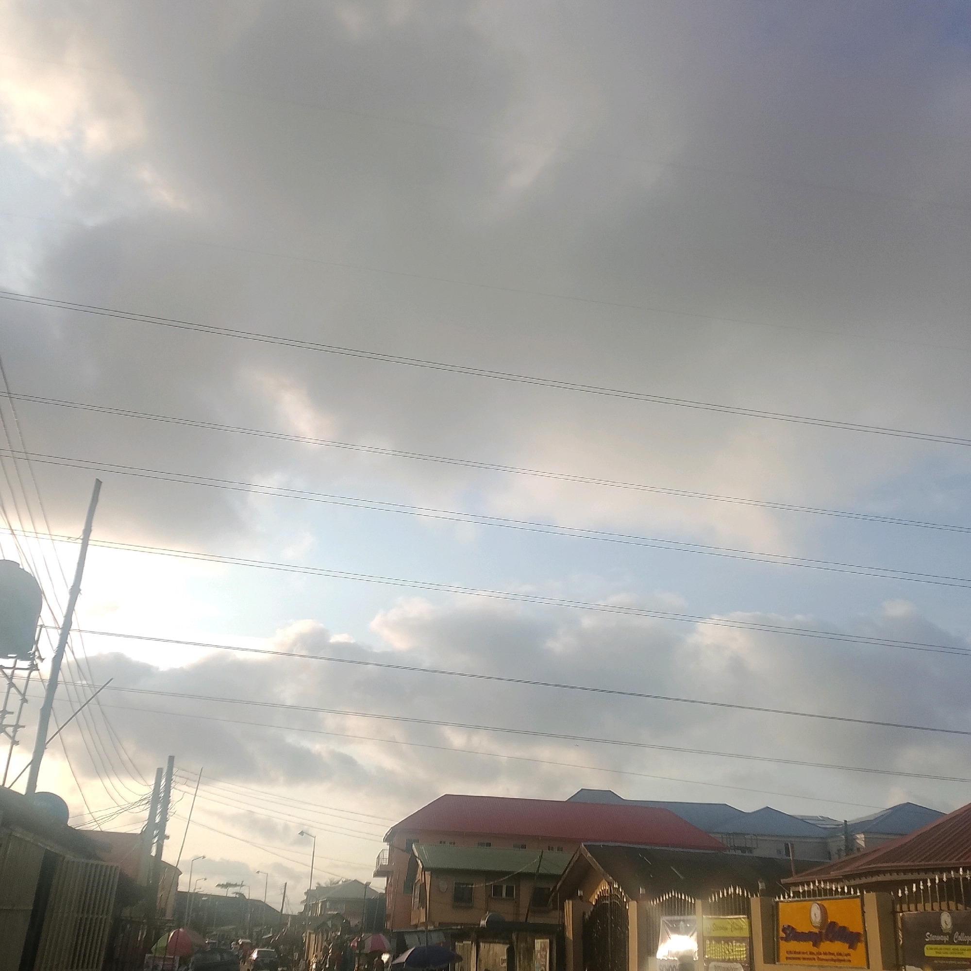 This image depicts a street scene in Ado, Nigeria, with a focus on the overhead power lines and a cloudy sky. The foreground shows the tops of buildings, some with red and green roofs, and a partial view of an outdoor market or street stalls. Electrical wires are strung across the frame, supported by utility poles, creating a dense network against the sky. The sky itself is dominated by heavy clouds with patches of sunlight breaking through, suggesting either early morning or late afternoon light. The overall impression is of an urban environment with active infrastructure and a somewhat overcast weather condition.