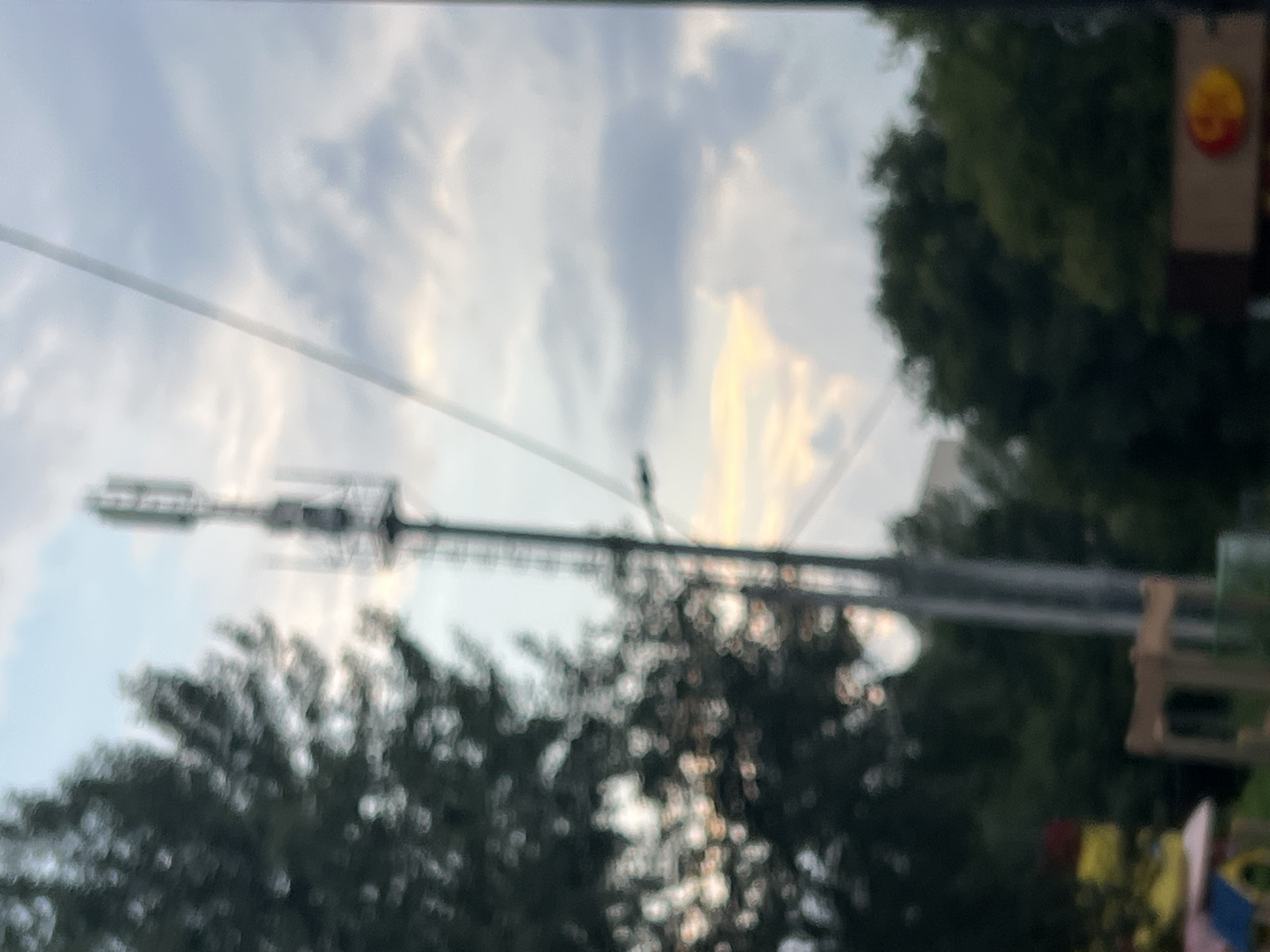 The image shows a blurry outdoor scene with a telecommunications tower dominating the center, silhouetted against a cloudy sky with patches of sunlight. To the left, the dark, out-of-focus foliage of a tree is visible. In the lower portion of the image, a brightly colored structure, possibly part of a playground, is partially obscured and out of focus. Further to the right and lower, there is a dark sign with a circular logo featuring red and yellow elements. The overall impression is that of a street or park area in Ulyanovsk, Russia, captured with a shallow depth of field, rendering most details soft and indistinct.