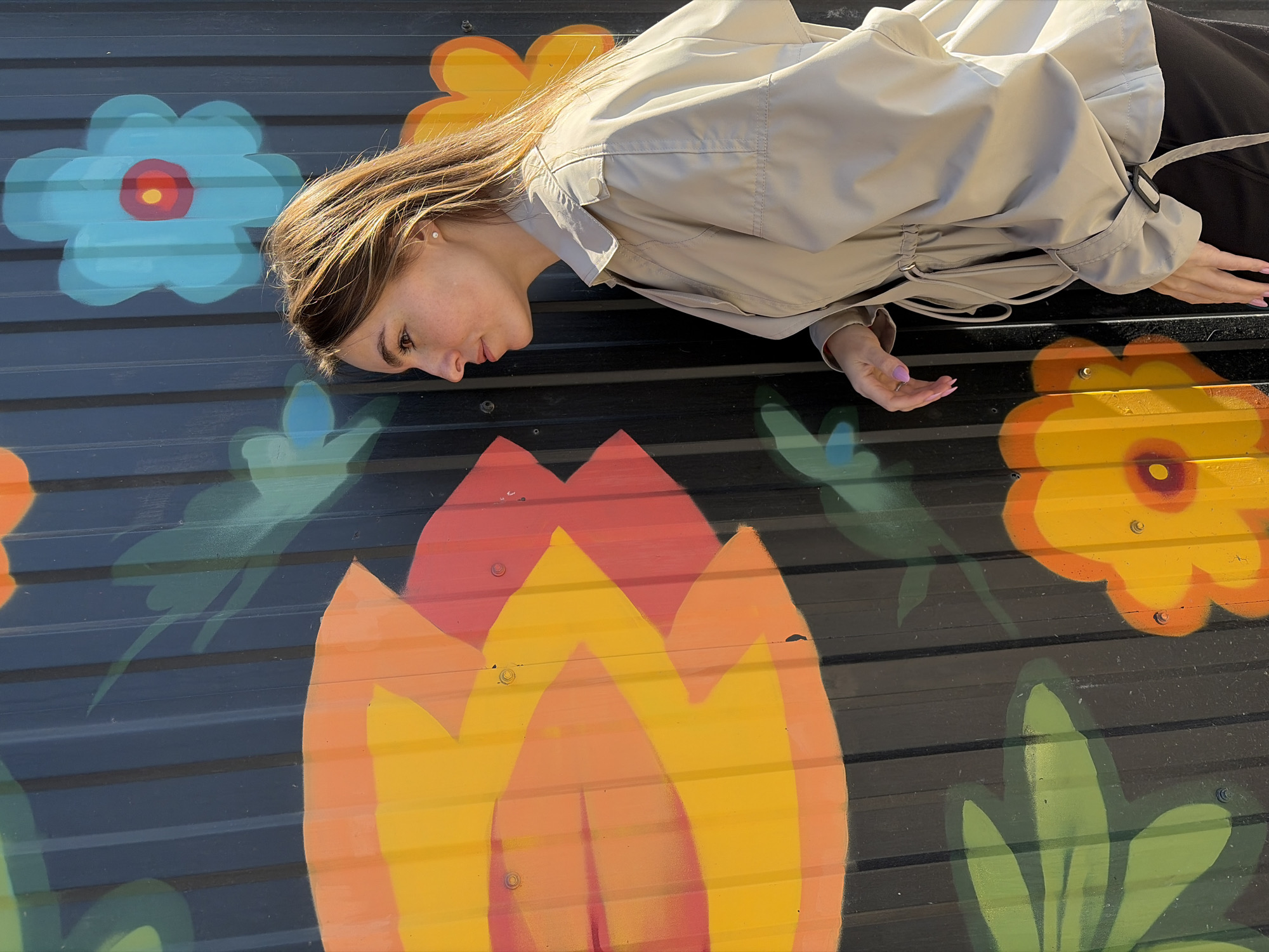 A young woman in a light tan trench coat is lying on a colorful mural in Izhevsk, Russia. The mural depicts oversized, stylized flowers in shades of blue, orange, and yellow against a dark background of corrugated metal. The woman is positioned horizontally, with her head near a blue flower and her body extending towards an orange and yellow flower. Her hands rest lightly on the mural's surface. Her hair is brown, and she is looking down. The overall impression is a casual, relaxed pose against a vibrant, urban backdrop.