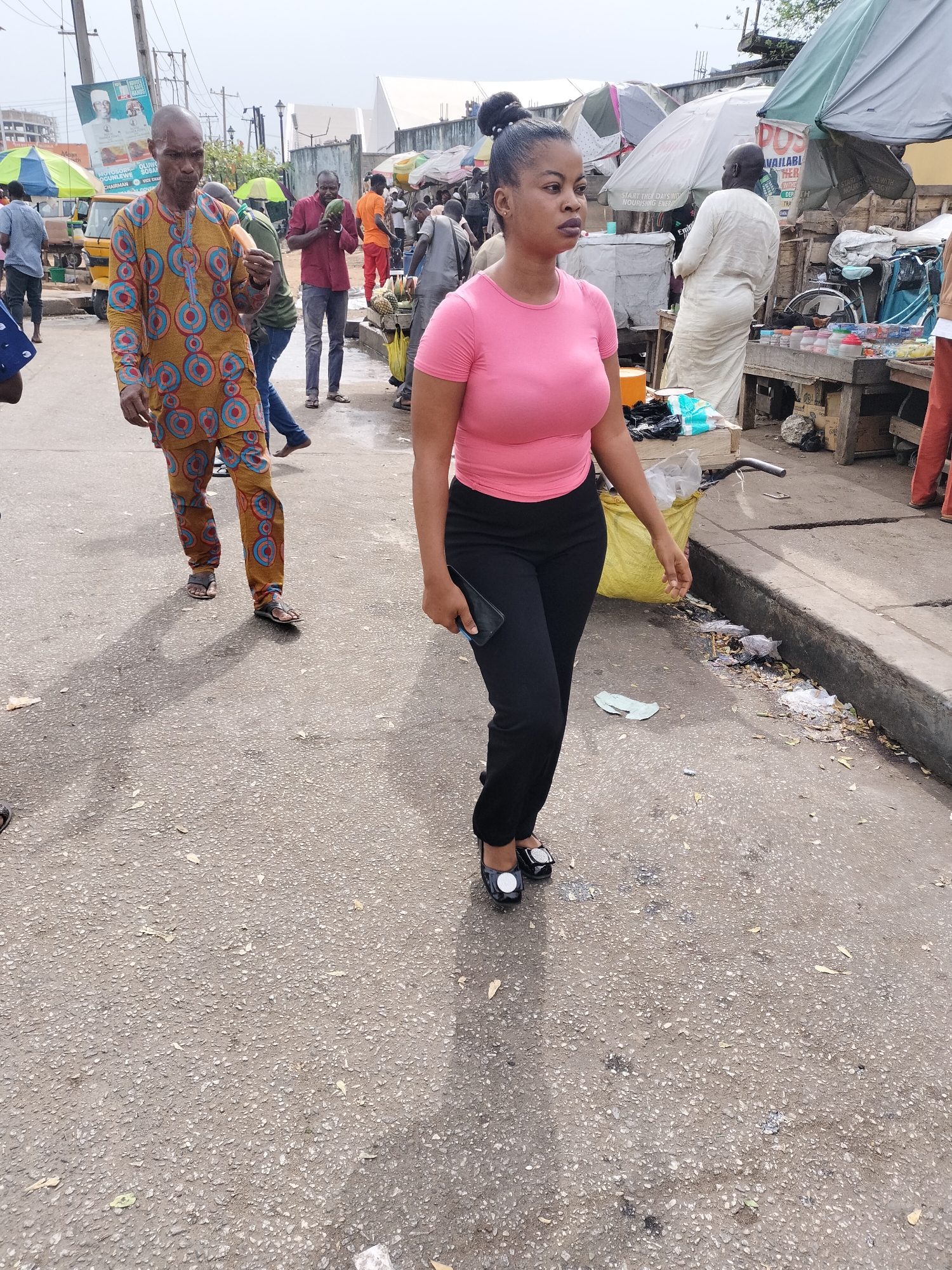 The image depicts a street scene in Lagos, Nigeria, capturing a moment of everyday life in a bustling environment.
In the foreground, a woman wearing a pink top and black trousers is walking towards the right of the frame, holding a cell phone. A man in a brightly patterned traditional Yoruba attire walks in the opposite direction.
Numerous other figures occupy the background, including a man in a white robe, likely a Muslim cleric, near a table laden with goods for sale. Several other individuals are scattered throughout the scene, engaged in various activities.
The setting appears to be a marketplace or a busy commercial area. Various vendors are set up along the side of the street. The road surface is concrete, with noticeable signs of wear and tear. Litter and debris line the curb, adding to the scene's grittiness.
Signs of commerce are evident in the image, with vendors displaying their wares under umbrellas and canopies. The overall impression is of a lively, crowded urban environment.