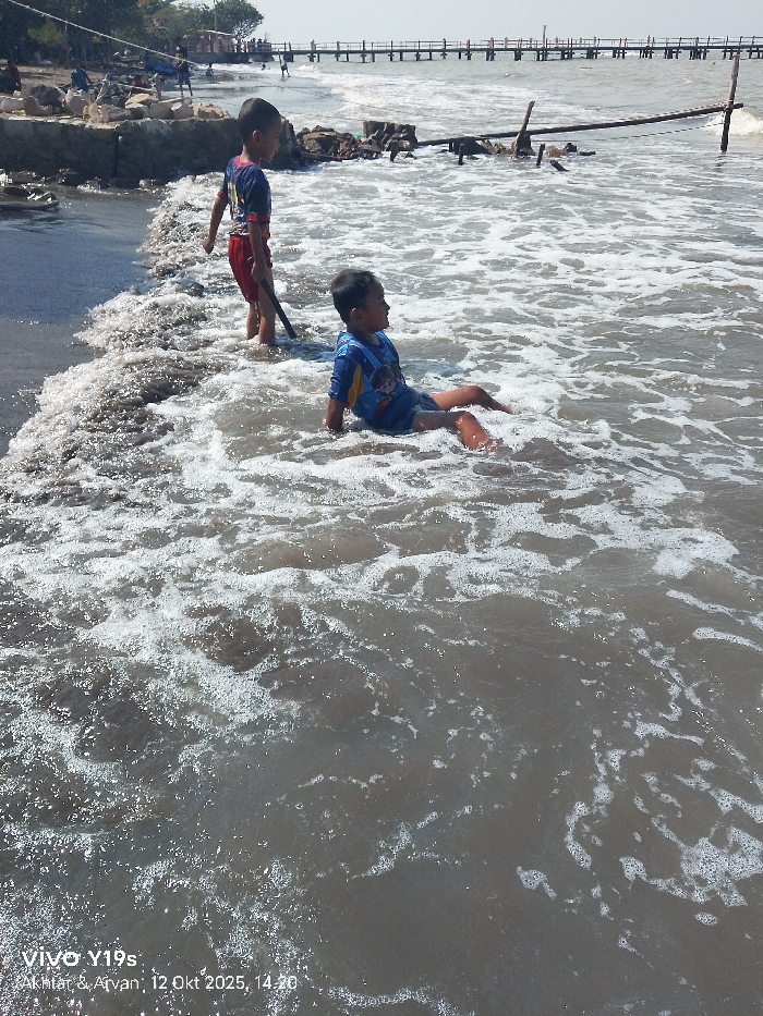 An outdoor scene is captured at 14:20 on October 12, 2025, in a coastal area of Limbangan, Indonesia. The image shows two young Asian male children playing in shallow, murky seawater under a bright, clear sky. In the mid-ground, one child, wearing a dark-colored short-sleeved shirt with red accents and red shorts, stands in the breaking waves, holding a dark stick. He is positioned facing right, looking towards the other child. To his right and closer to the foreground, a second child, dressed in a blue short-sleeved shirt with a yellow graphic design and dark shorts, sits submerged from the waist down in the water, with his legs extended forward, facing left. The water is brownish-grey with white foam from small waves. The immediate shoreline to the left consists of dark, wet sand backed by a low, rocky embankment with scattered debris. In the background, a long wooden pier extends horizontally into the sea, supported by numerous posts, with several indistinct human figures visible on its structure. Additional broken wooden posts protrude from the water closer to the pier.