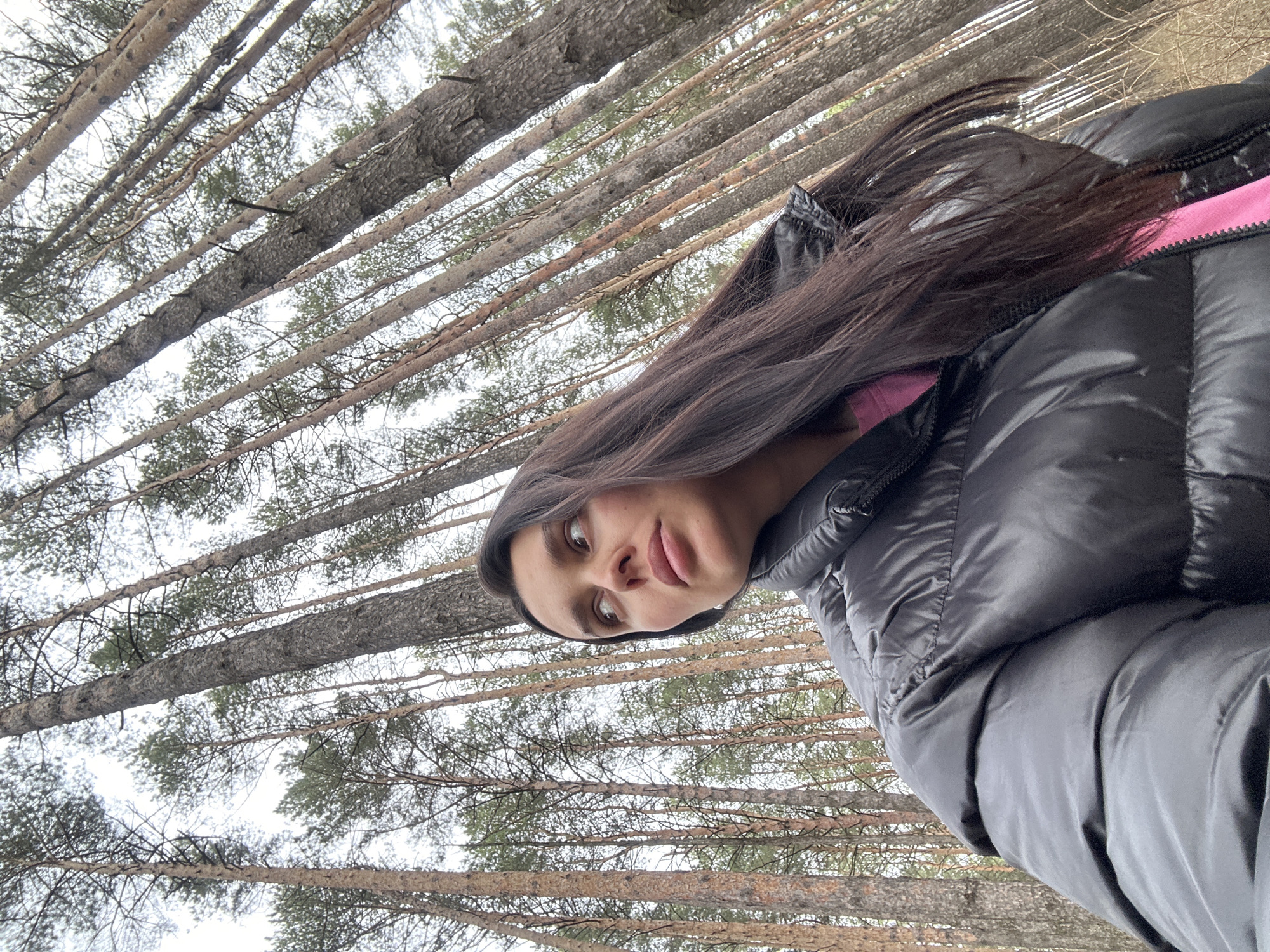 A person with long, dark hair, wearing a black puffer jacket with a hint of a pink shirt underneath, is positioned in the foreground. The backdrop consists of numerous tall trees, most likely pine trees, extending upwards towards a muted sky. The perspective is from below, looking up through the trees. The location is Chaikovsky, Russia, based on the likely context or additional information that is not part of the provided image.