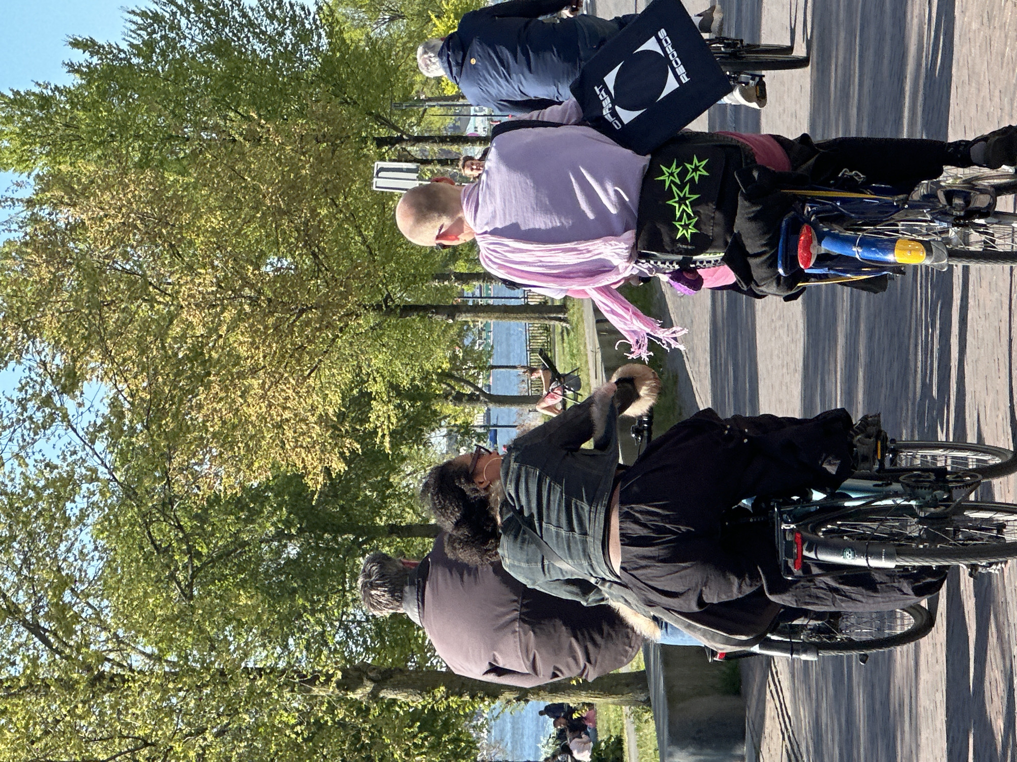 The image captures a group of people cycling along a paved path next to a body of water, likely a canal or river, under a clear blue sky. The scene is bright and sunny, with lush green trees lining the path, suggesting a warm, pleasant day in spring or summer.
In the foreground, two individuals are seen from behind, riding bicycles. The person closest to the viewer wears a dark brown or grey jacket. Ahead of them is another person with dark, curly hair, wearing a dark, possibly denim, jacket with fur trim on the collar and cuffs, and dark pants. Their bicycle is light blue/grey.
Further along the path, two more cyclists are visible. One person, bald, wears a light lavender shirt and a pink scarf draped around their neck, with a black bag on their bike featuring three bright green stars. Their bike has a red taillight and a yellow reflector. Behind them, another person in a dark blue jacket is also on a bicycle, carrying a black tote bag over their shoulder. The text "RECORDS" is visible on this bag, along with a stylized geometric logo (a circle with a smaller circle and a triangle inside).
The path itself is paved with light-colored material, showing long shadows cast by the trees and cyclists, indicating the sun is relatively low in the sky, perhaps late morning or afternoon. In the background, across the body of water, more trees and hints of buildings can be seen. The overall impression is a leisurely ride in an urban park setting, consistent with locations like Amsterdam.
