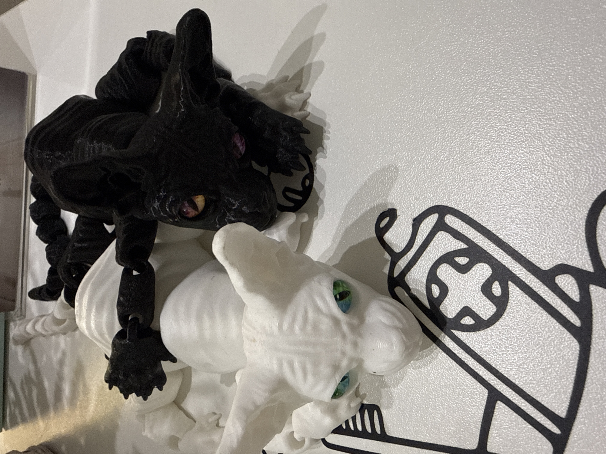 The image displays two stylized cat figurines on a textured white surface. The figurine on the left is white, with large pointed ears and vibrant green eyes. The figurine on the right is black, with orange and pink eyes, and its body is intertwined with the white figurine, suggesting a dynamic pose. A black, chain-like object connects the two figures. The surface appears to be a table or countertop with a black graphic design featuring geometric shapes, possibly representing a game controller. The lighting suggests an indoor environment. No geographical location or context beyond the objects themselves is discernible from the image.