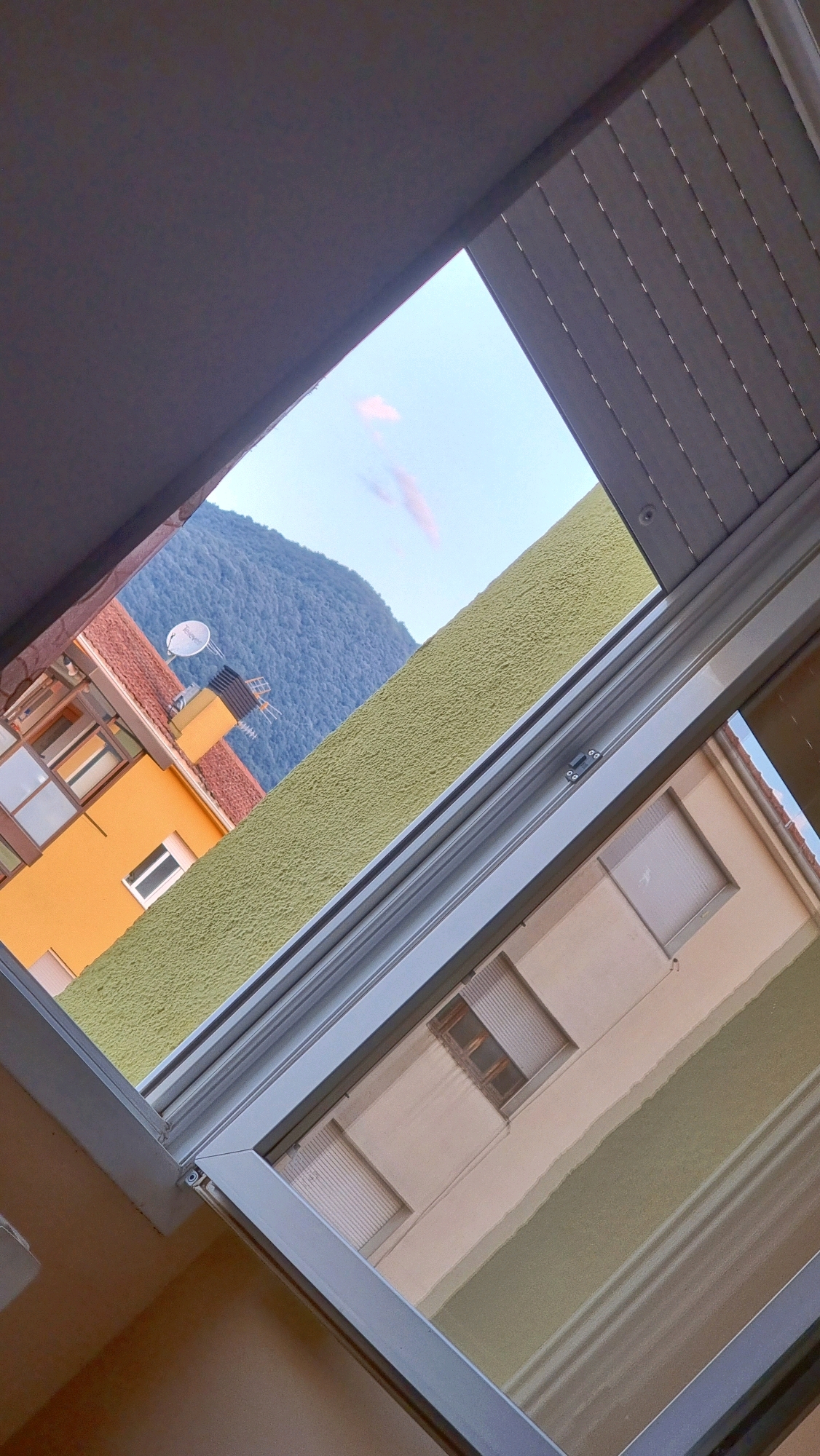 An eye-level, full, angled shot shows a view through a partially open window with a beige frame and a white trim. The window overlooks a light green textured wall and a blue mountain in the background. A yellow building with a red-tiled roof is visible on the left side of the frame, with a satellite dish and antenna on its roof. Reflections in the window show a light brown wall and a beige building with closed shutters. The scene appears to be a residential area in Sant Joan Les Fonts, Spain, taken on a clear day.