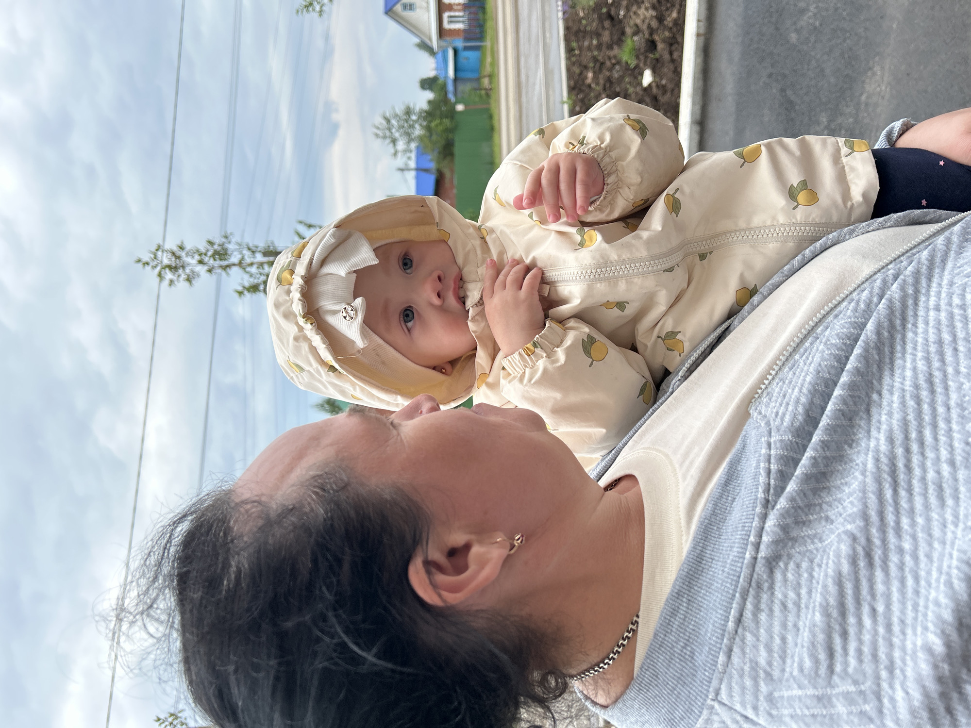 The image, oriented in portrait, captures a heartwarming interaction between an adult and a baby in an outdoor setting. In the foreground, an adult woman with dark, possibly greying, hair is visible from the shoulder up, facing right and looking towards the child. She wears a light-colored top underneath a light blue jacket with a subtle textured pattern and a silver chain around her neck. A small gold hoop earring is visible on her ear.
In the mid-ground, a baby or young child is positioned, looking upwards and slightly to the left, with curious blue eyes. The child is dressed in a cream-colored hooded jumpsuit featuring a repeating pattern of yellow lemons with green leaves. A white bow, possibly with a faint logo, adorns the hood. The child's fingers are near their mouth. Beneath the jumpsuit, dark blue leggings with small pink star patterns are visible.
The background shows an overcast or cloudy sky with visible power lines. Beyond, there are green trees and distant houses with distinctive blue and red roofs, suggesting a rural or suburban environment typical of locations like Азинский, Russia. The overall impression is a daytime scene under a soft, natural light, highlighting the tender moment between the subjects. No legible text is clearly visible.