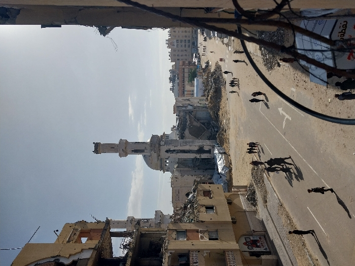 This image depicts a devastated urban landscape in the city of Khan Yunis, Palestinian Territory, viewed from an elevated position under a bright, clear sky.
In the mid-ground, a large mosque with a tall, slender minaret and a dome stands prominently. The main structure of the mosque is heavily damaged, with large sections of its facade and dome partially collapsed, exposing internal debris. The minaret itself appears mostly intact at its upper section.
To the left of the mosque, a multi-story building is severely damaged, with significant portions of its outer walls missing, revealing internal floors and rubble. Piles of debris are visible around its base. Additional damaged structures are present in the distant background.
A paved road occupies the central and left-foreground, featuring white directional arrows painted on its surface: a left-turn arrow and a combined straight/right-turn arrow. The areas immediately adjacent to the road and extending towards the background are unpaved, consisting of dirt, sand, and extensive piles of building rubble.
Approximately 20-30 individuals are present on the street and the surrounding dirt areas. They are primarily adults, depicted walking either singly or in small groups. Their attire is civilian. Shadows are cast distinctly on the ground due to the overhead sun.
The scene is framed on the immediate right by a dark, curved metallic element, possibly a balcony railing or wires, partially obscuring the view. The sky is bright and largely clear, with a visible light glare in the upper right quadrant.