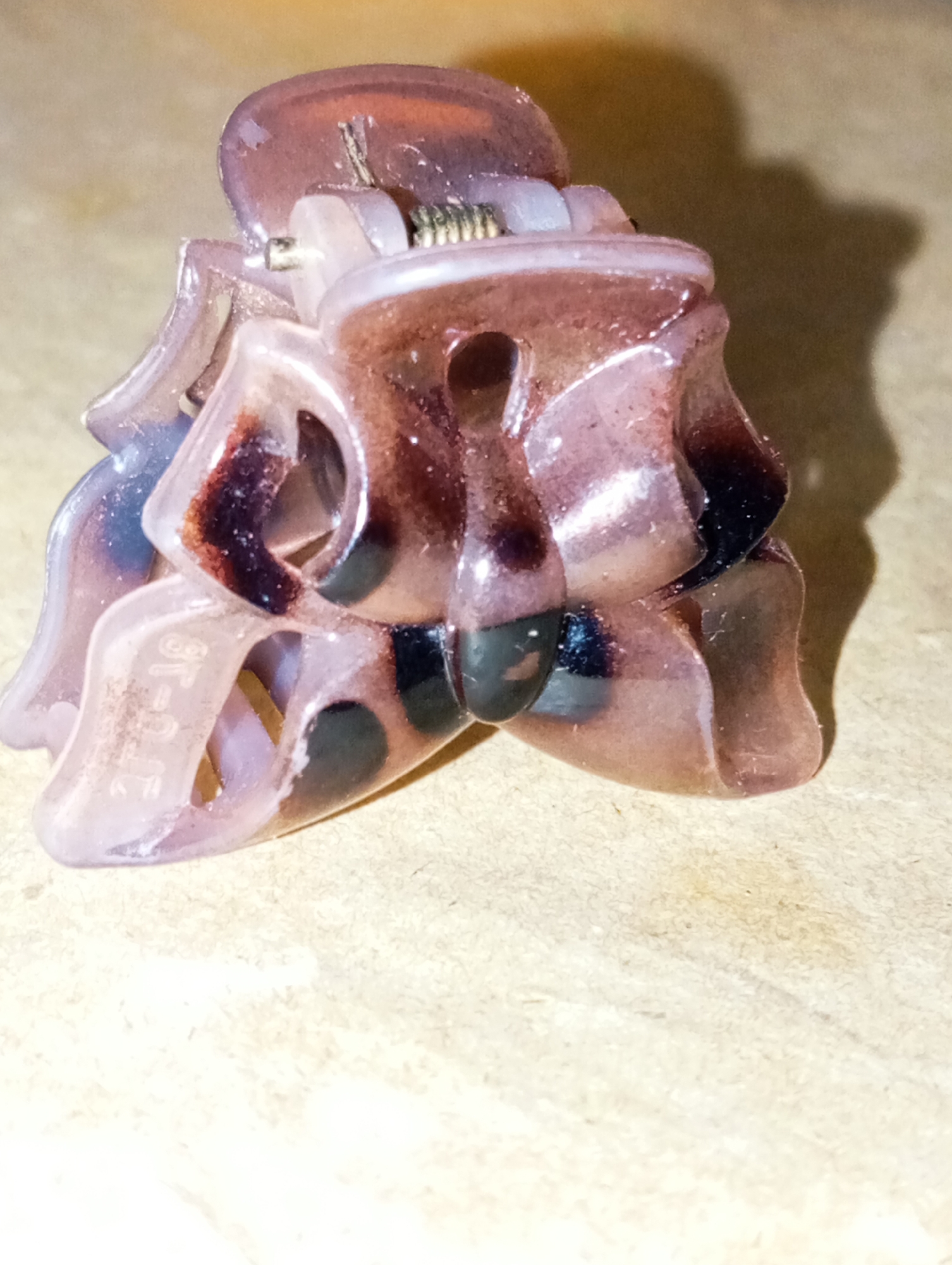 A close-up, eye-level shot features a single plastic hair claw clip. The clip is primarily a translucent pinkish-purple with dark brown and black marbling and spots. It is shaped like a flower, with four distinct petal-like sections that come together at the top to form the gripping mechanism. A metal spring is visible in the center, connecting the two halves of the clip. The clip is lying on a textured, beige surface that appears to be cardboard or wood. The lighting creates a shadow of the clip on the surface to the right. There are no people visible in the image, and the scene is not recognizable as being in any specific city or location.