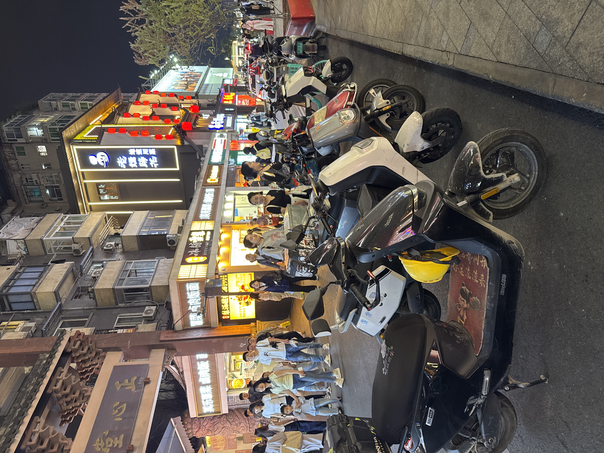 Here is a detailed description of the image:
**General Scene:** The image captures a bustling night scene in Jinjiang District, China. A narrow street is lined with shops and buildings, with electric scooters parked along the side. A large group of people is walking on the street.
**Buildings and Shops:** The buildings are multistoried with traditional Chinese architectural elements. Shops have bright neon signs in Chinese characters. Red lanterns are hung above some of the buildings. The signs include "其乐薯菜卤肉饭" and "老燻蹄花."
**Vehicles:** Numerous electric scooters are parked along the right side of the street. The scooters are of various makes and models, mostly white and black in color. Some have personalized seat pads or decals.
**People:** A large group of people walks in a line along the road. Most of them seem to be wearing white t-shirts. A lot of the people are wearing masks.
**Overall Impression:** The image depicts a vibrant, modern urban environment in China, with a blend of traditional architecture and contemporary transportation. The crowd and bright lights suggest a lively evening atmosphere.