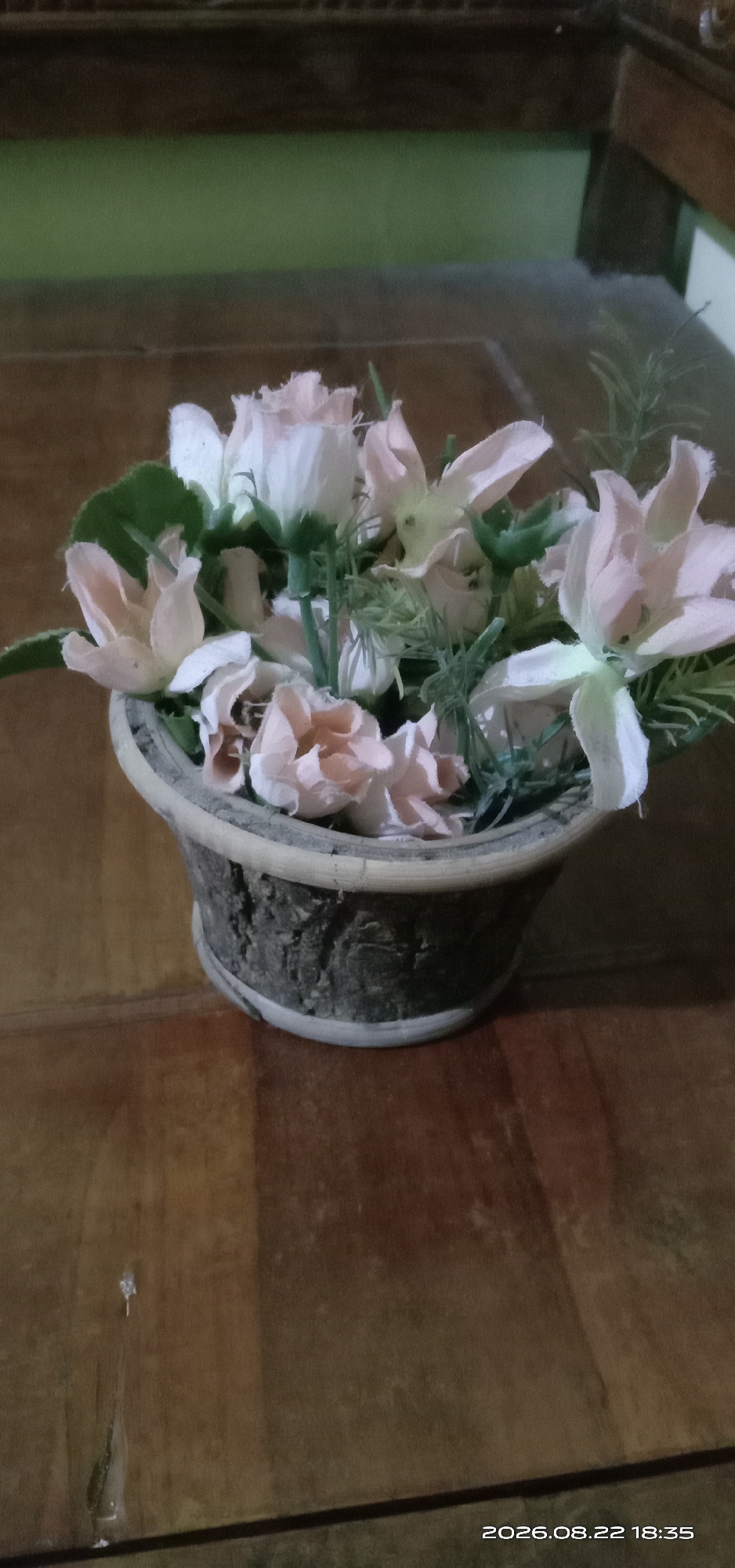 A floral arrangement of light pink and white artificial flowers sits in a textured, dark brown pot on a wooden surface. The pot has a lighter rim. In the background, a green wall is visible above a dark wooden structure. The timestamp "2026.08.22 18:35" is overlaid on the lower right corner of the image. The scene is indoors, likely a home or office. There are no people present. The image is taken in Pakong, Indonesia.