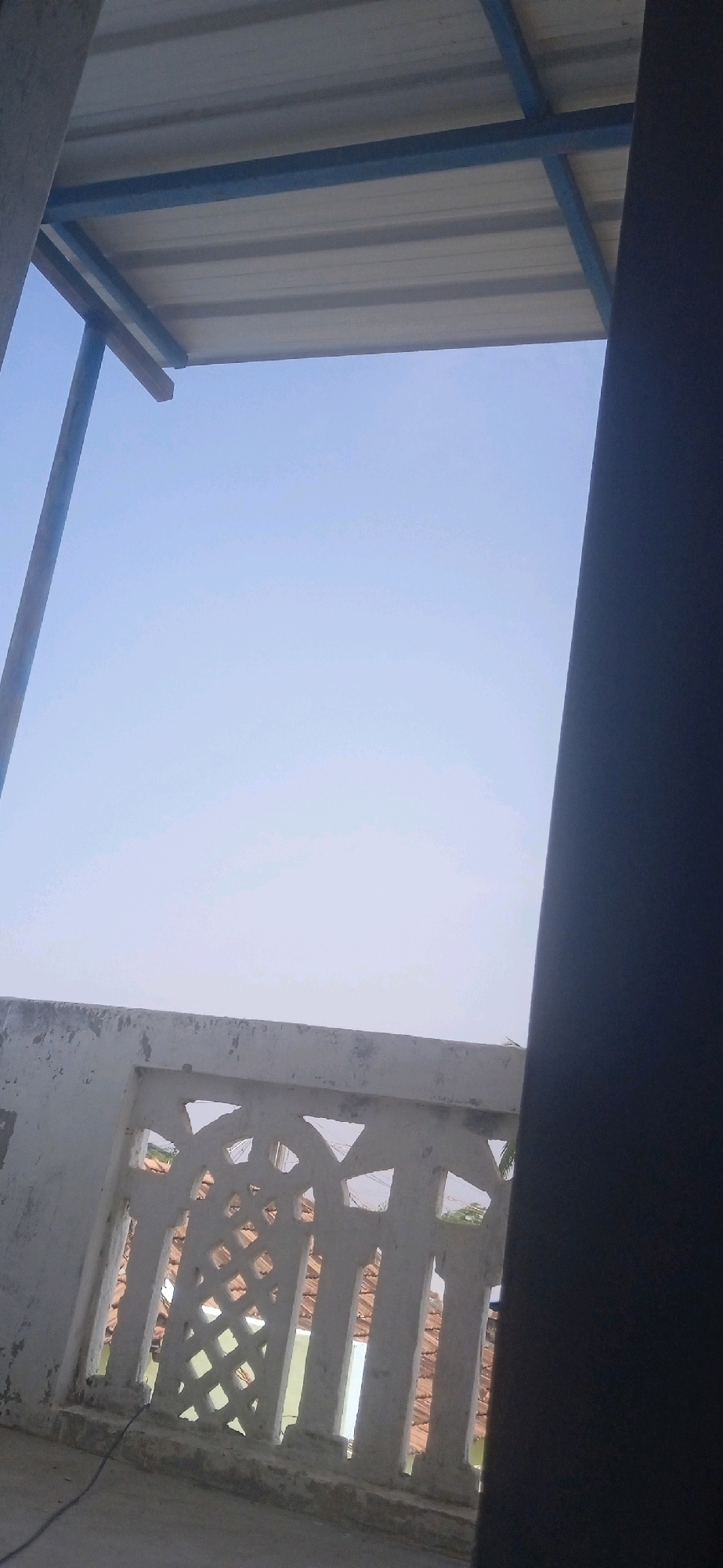 The image depicts a view from what appears to be a balcony or an elevated structure, looking upwards towards a clear sky and downwards at a decorative concrete railing. The railing has a patterned design with triangular and diamond-shaped openings. Beyond the railing, roofs of buildings with terracotta tiles are visible, suggesting an urban or semi-urban environment. The sky is a pale blue, indicating daytime. In the upper part of the image, a section of a corrugated metal roof supported by metal beams is visible, casting a shadow. A dark vertical structure, possibly a wall or pillar, occupies the right side of the frame, partially obscuring the view. A thin black cable is visible on the ground near the railing. The scene is consistent with a residential area in Anaipady, India. There are no people visible in the image.