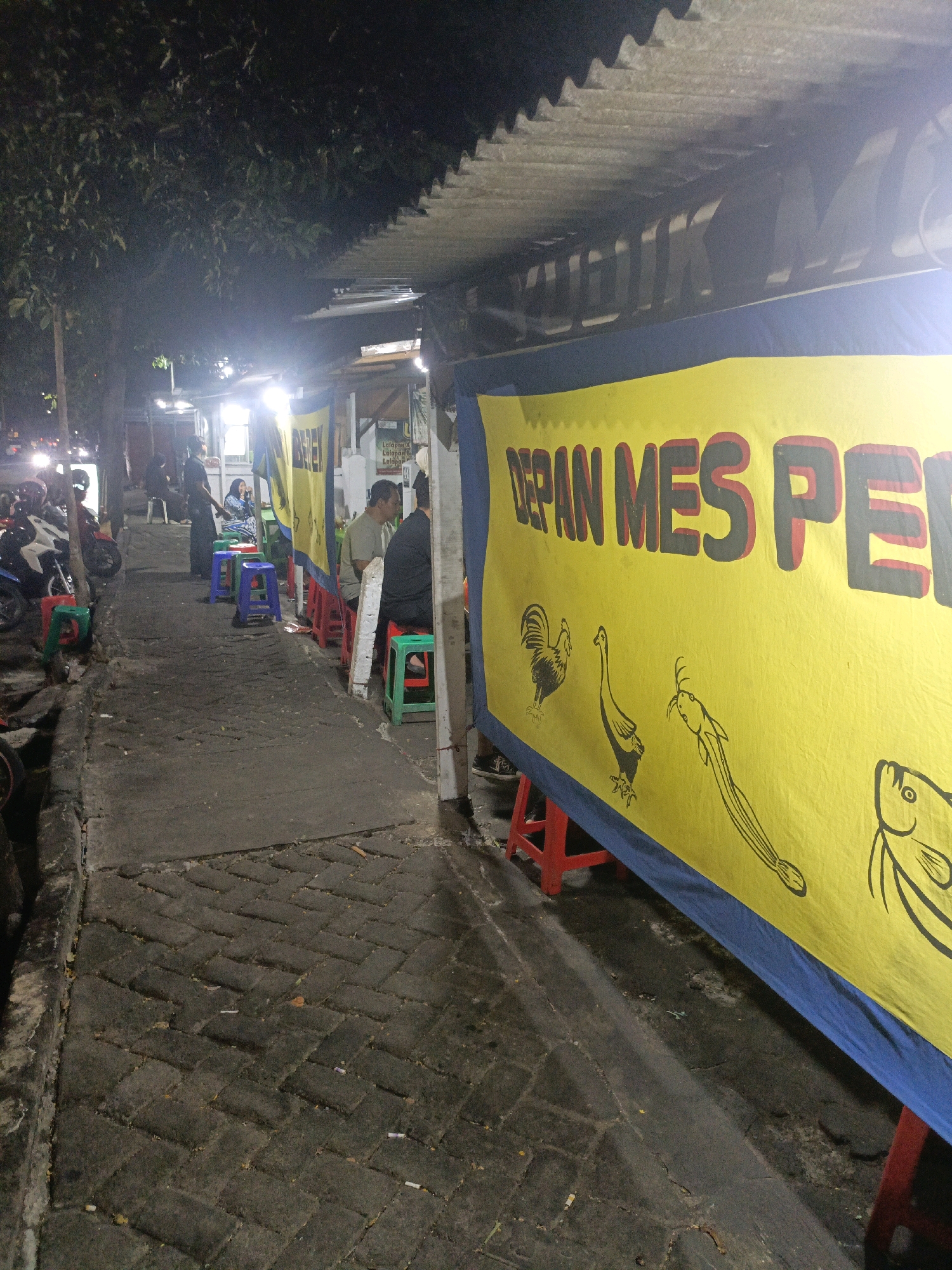 The image depicts a nighttime scene on a sidewalk in Probolinggo, Indonesia, likely a food stall area. A prominent yellow banner with blue trim stretches across the foreground, displaying large, dark text that reads "DEPAN MES PE..." and features simple black line drawings of a rooster, a bird resembling a duck or goose, a catfish, and a fish. Red and green plastic stools are scattered along the sidewalk, some occupied by patrons. To the left, a street with parked motorcycles and passing vehicle headlights suggests proximity to a road. The environment is dimly lit by artificial lights, casting shadows and highlighting the textured brick paving of the sidewalk. Several individuals are visible, some seated at the stools, others standing, engaged in what appears to be dining or socializing at the outdoor establishment. The overall atmosphere is that of a casual, late-night eatery.